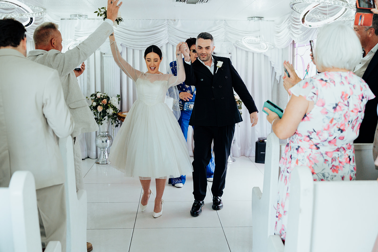 Smiling couple exits The Little White Wedding Chapel hand in hand as guests cheer