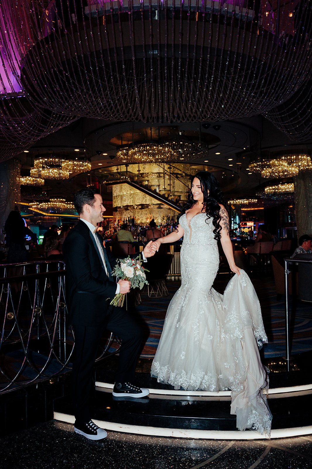 Bride in a lace wedding gown holds her train as she walks down a grand staircase while her groom kneels, offering her flowers. The setting is illuminated with glittering chandeliers and vibrant purple lighting.