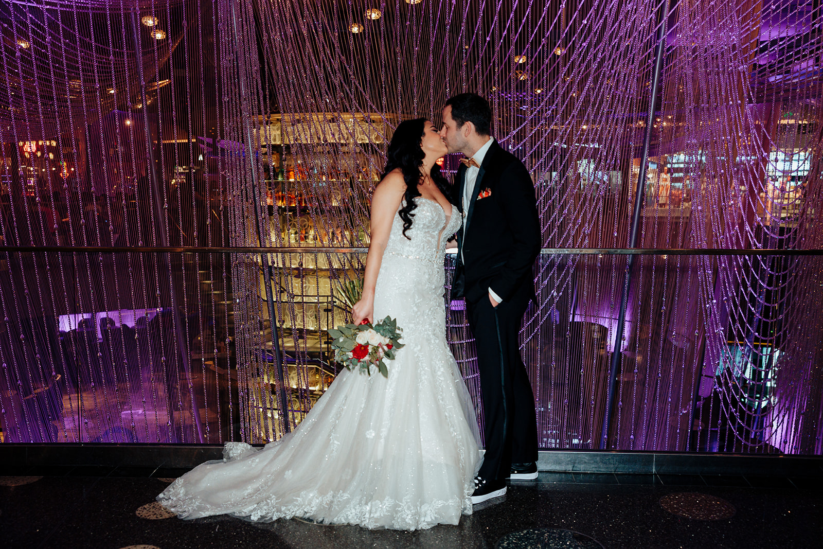 Bride and groom kiss on a balcony surrounded by shimmering purple lights at a Las Vegas wedding venue, holding a bouquet of red and white flowers.