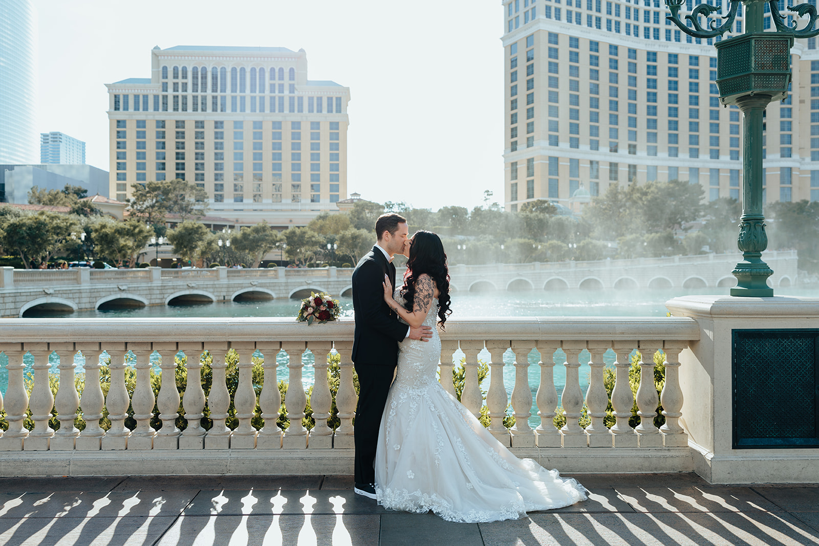 Bride and groom stand on a balcony in front of the Bellagio Fountain, sharing a romantic kiss while dressed in wedding attire.