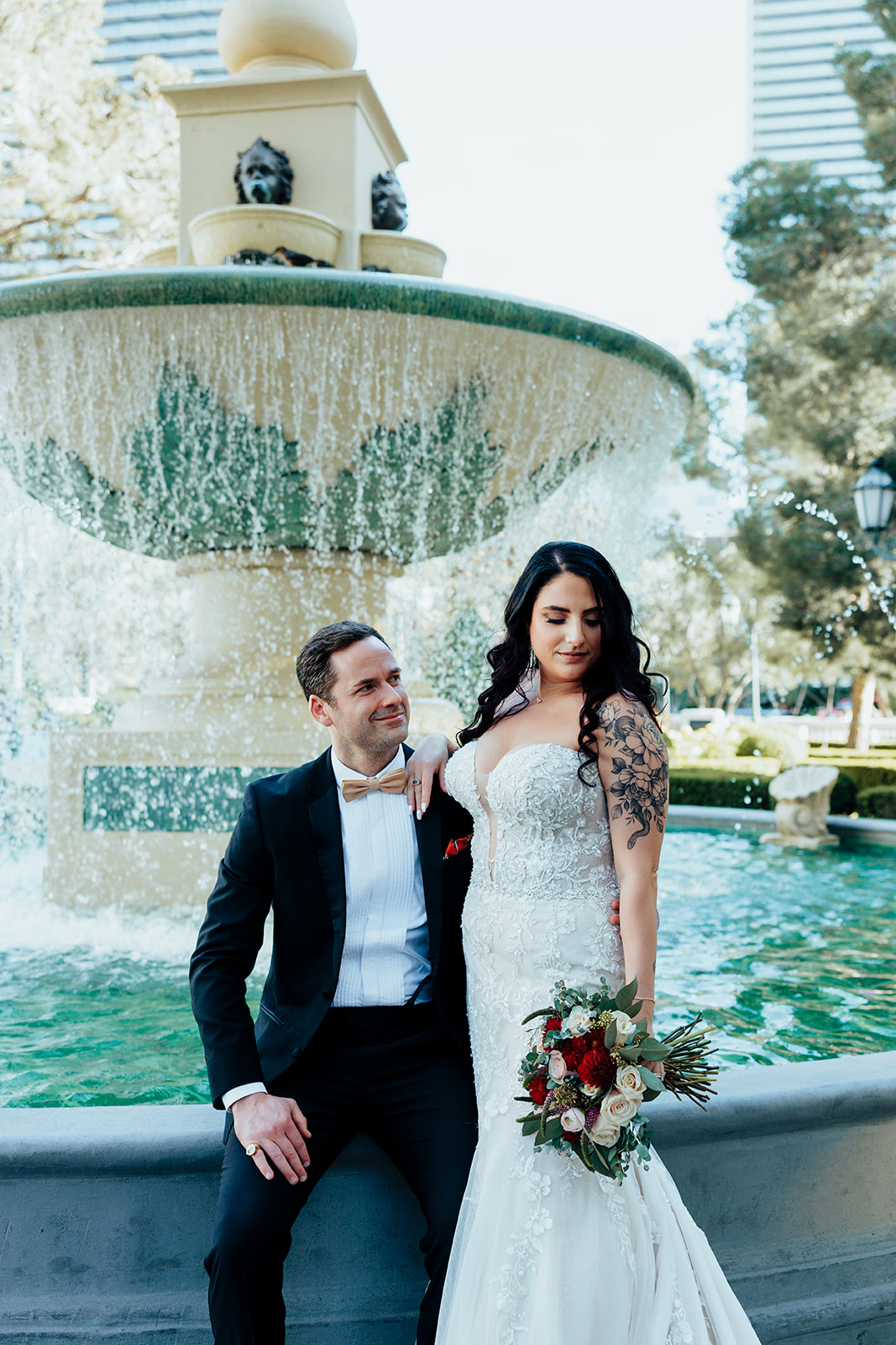 Bride and groom pose near a cascading fountain surrounded by lush greenery in a Las Vegas garden. The bride holds a red and white bouquet, while the groom gazes lovingly at her.