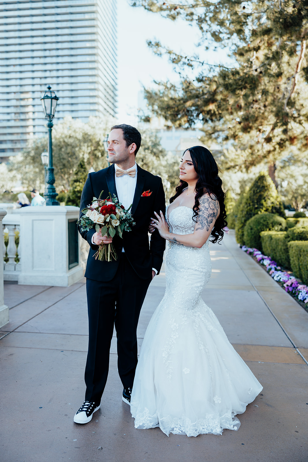 Bride and groom stroll along a flower-lined walkway in a Las Vegas garden. The bride holds a bouquet of roses while the groom looks thoughtfully ahead.