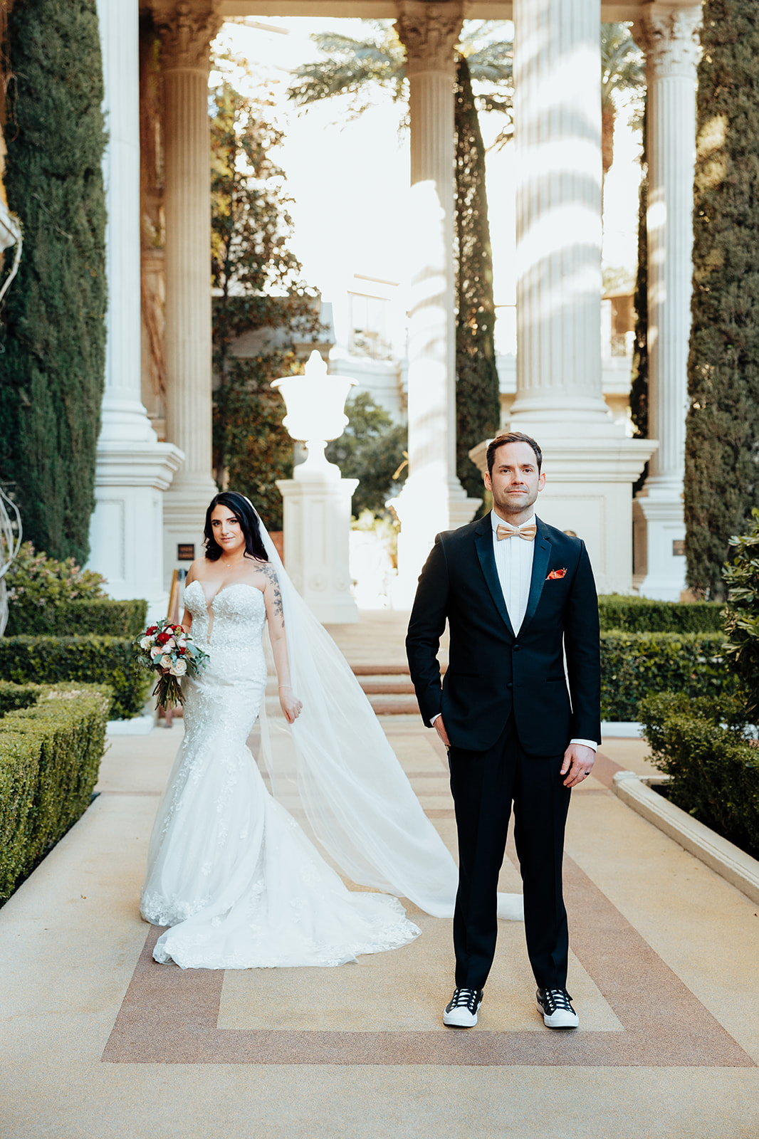 Groom standing confidently while bride walks behind with bouquet