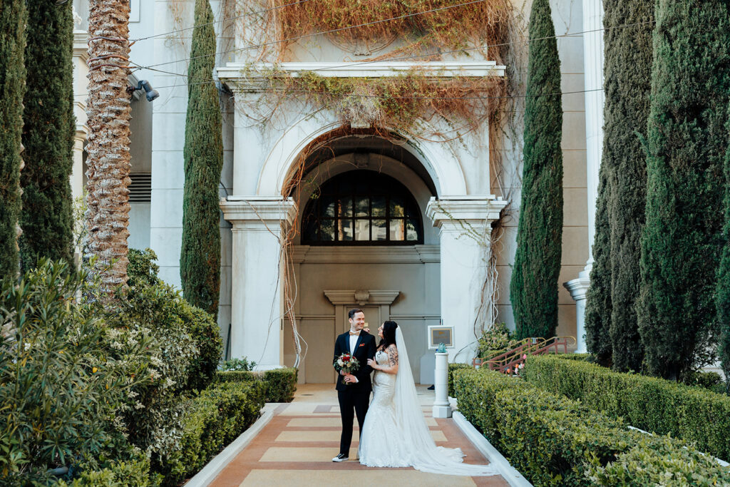 A bride in a lace wedding gown and long veil stands beside her groom in a tuxedo, holding a bouquet of red and white flowers. They are framed by lush greenery and an arched architectural backdrop.