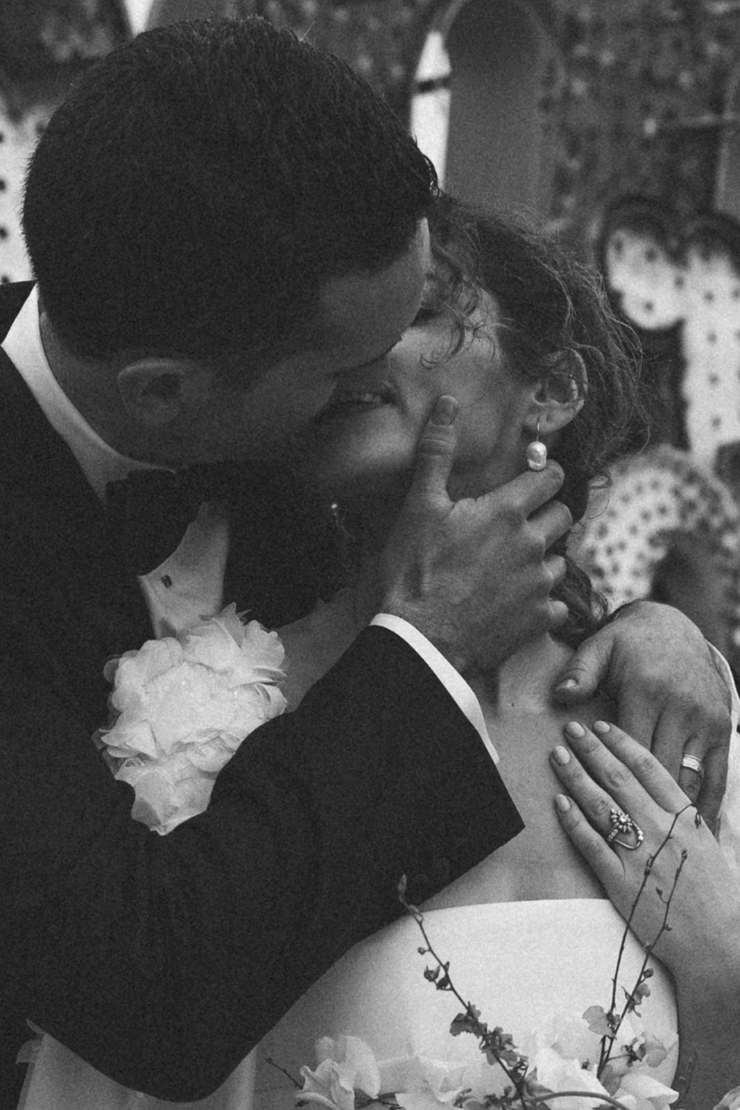 Black and white close-up of the groom gently holding the bride's face as they share an intimate kiss, with floral details and elegant wedding attire.