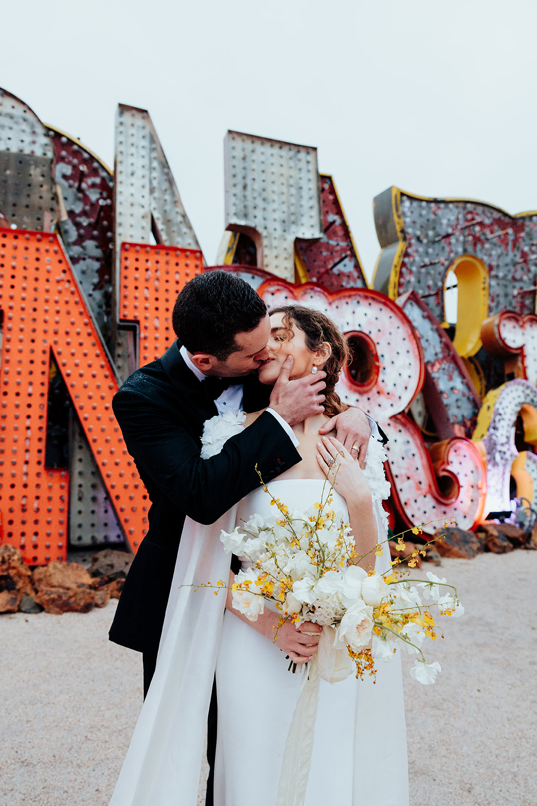 Bride and groom sharing a passionate kiss, with the groom holding the bride's face and the bride holding a bouquet of white and yellow flowers, against a vibrant backdrop of vintage neon signs.