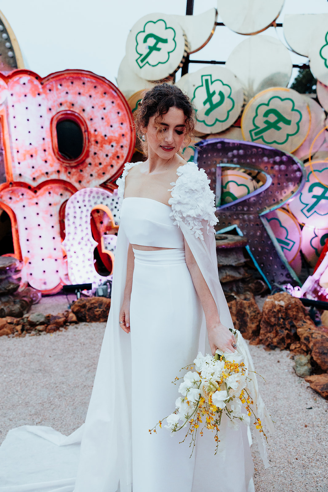 Bride wearing a white dress with floral detailing on the sleeves, holding a bouquet of white and yellow flowers, standing in front of colorful vintage neon signs at the Neon Museum.