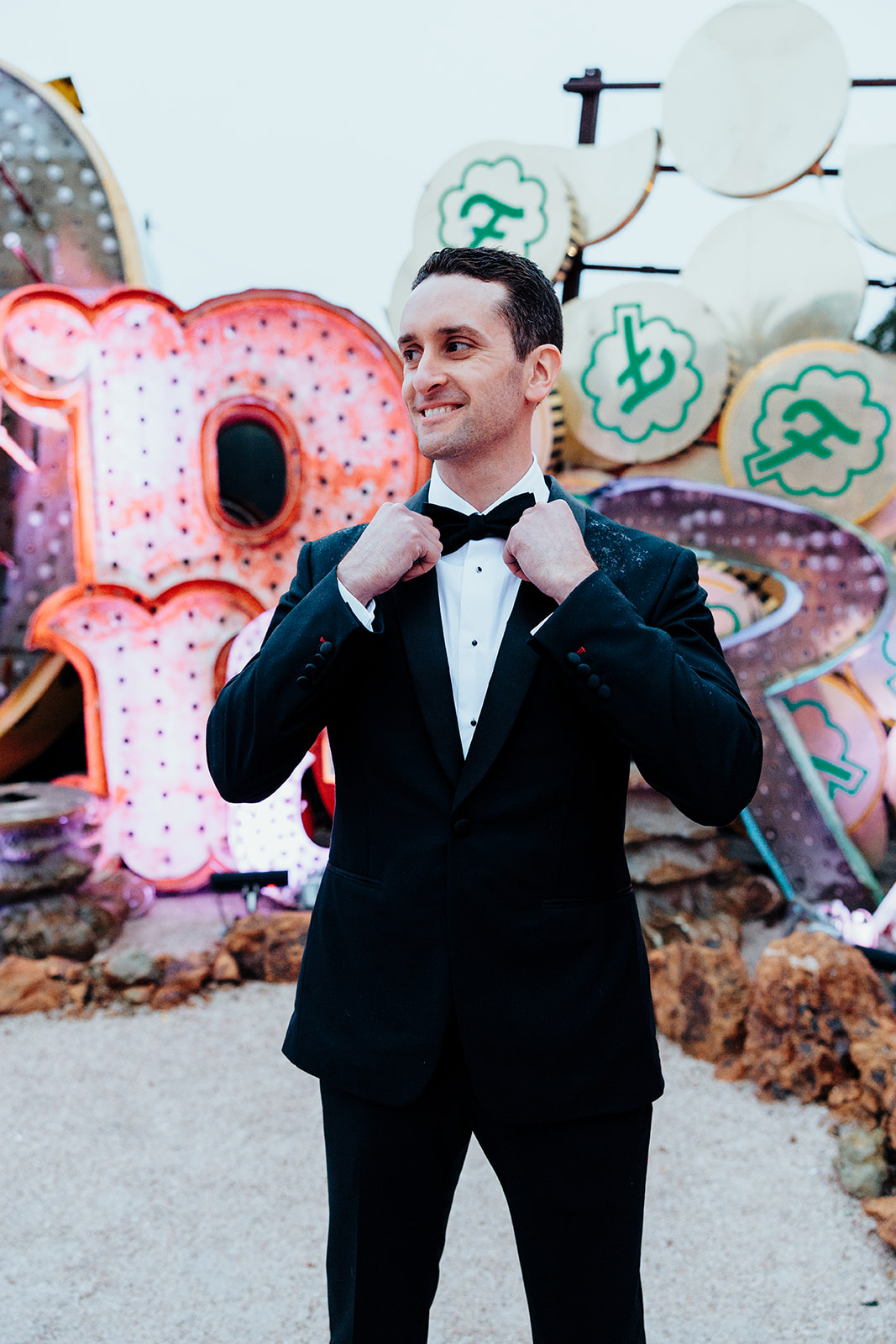 Groom in a black tuxedo adjusting his bow tie with a smile, standing in front of colorful vintage neon signs at the Neon Museum.