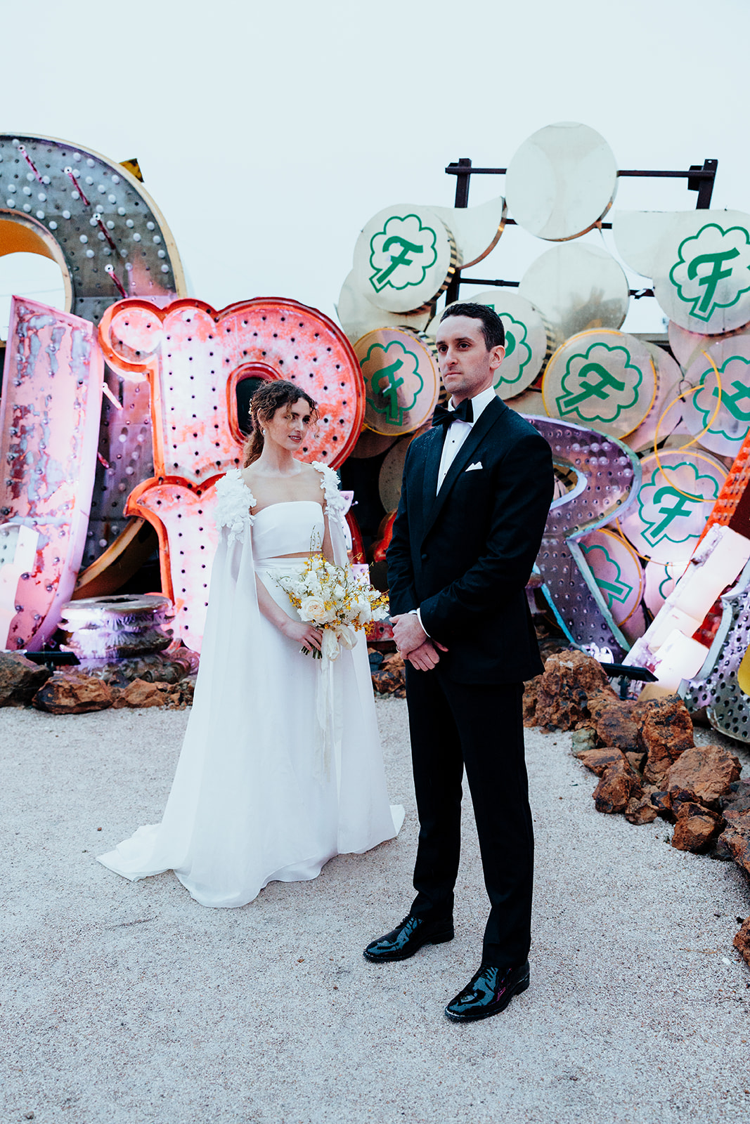 Bride in a white gown with a floral bouquet stands next to the groom in a black tuxedo, posing against a backdrop of colorful vintage neon signs.