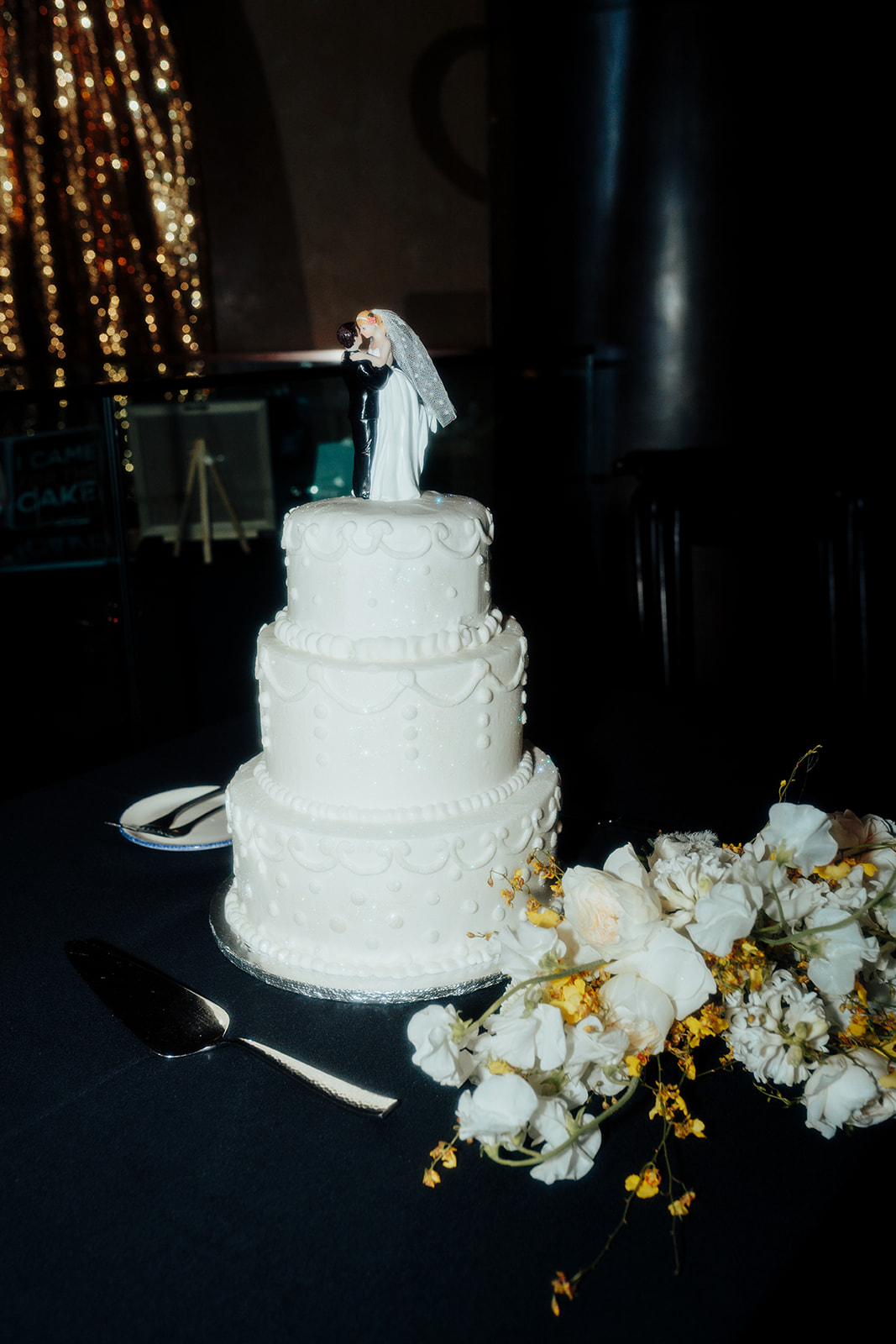 Close-up of a white wedding cake with a bride and groom topper, paired with a floral arrangement of white and yellow flowers at a Las Vegas wedding reception.