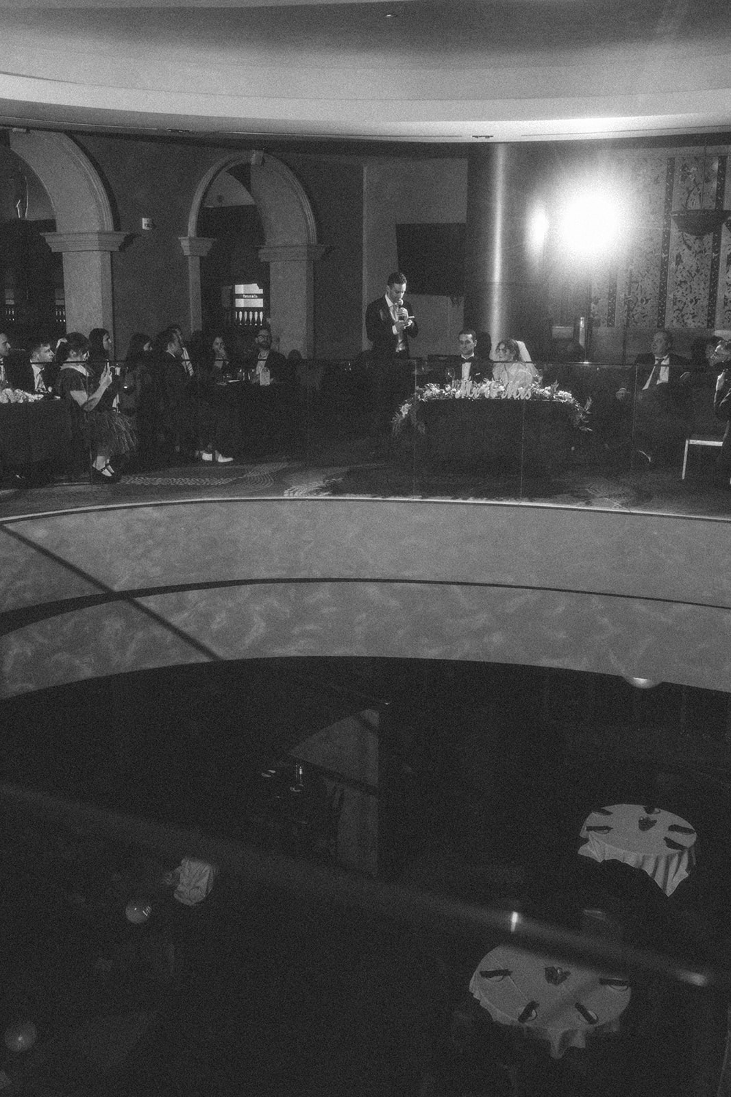 Black and white photo of the best man giving a speech during a Las Vegas wedding reception at Caesars Palace, with the bride and groom seated at the head table.