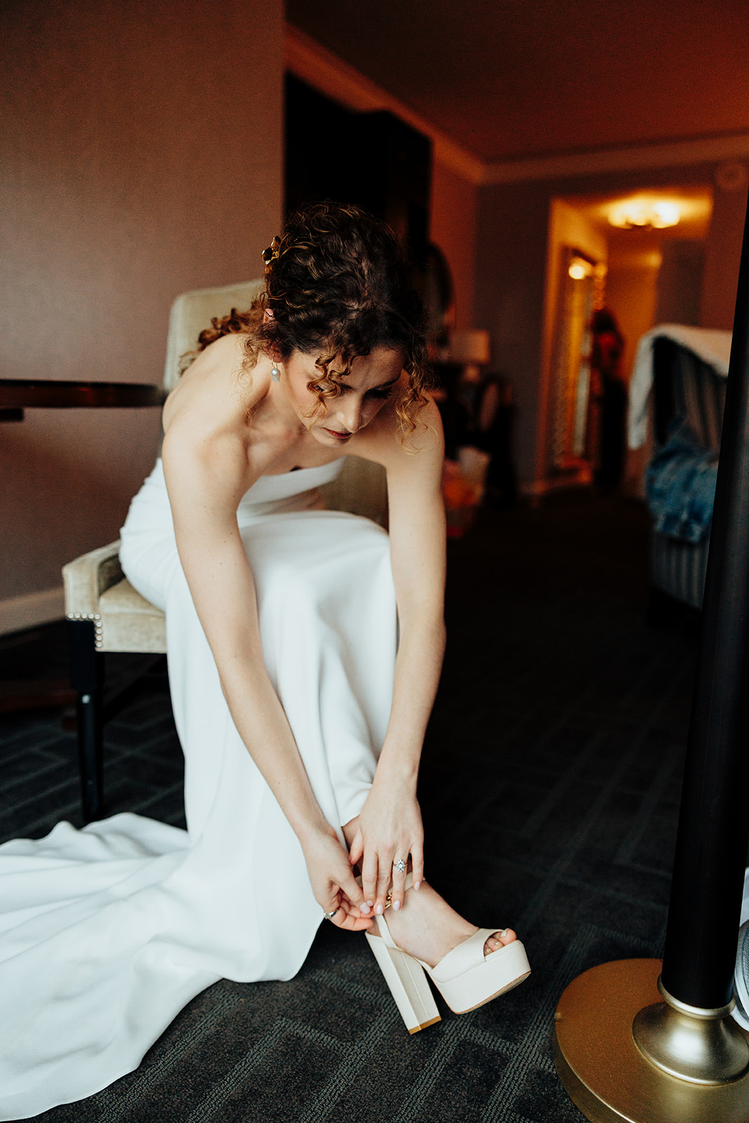 A Bride in Caesar's Palace in a white wedding gown adjusting her high-heeled shoe indoors before the ceremony, with a warm and elegant ambiance.