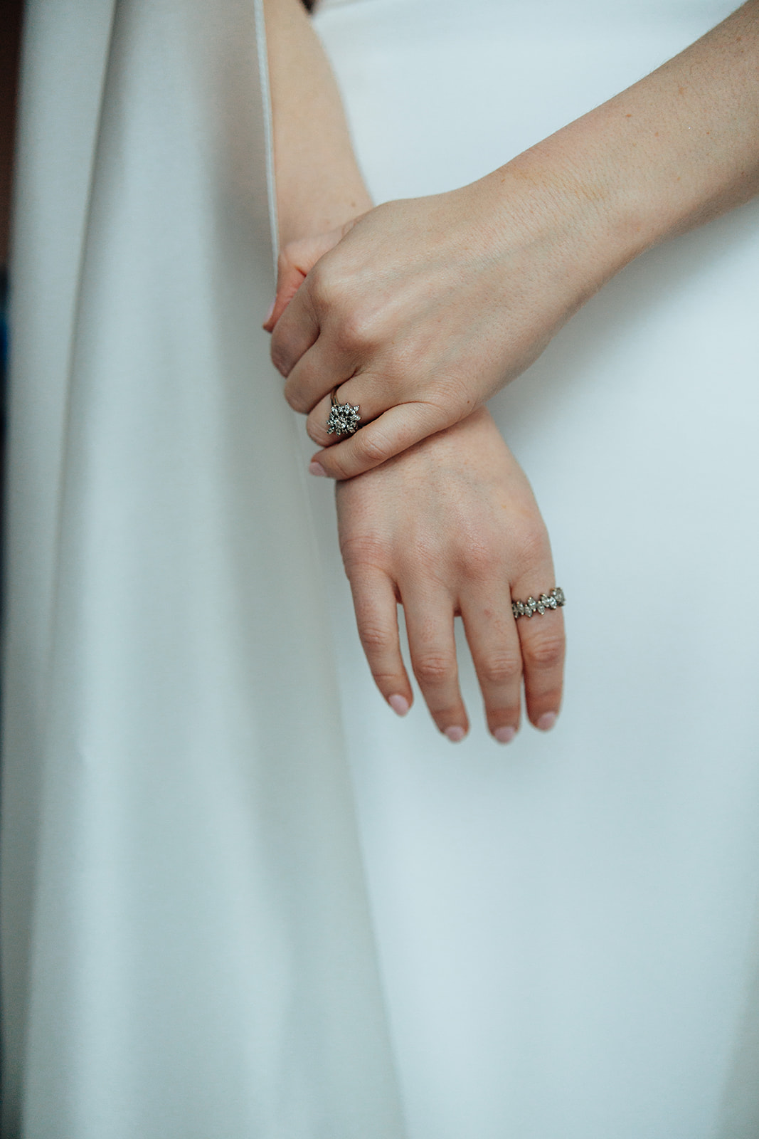 Close-up of bride's hands adorned with an engagement ring and wedding band resting against her white gown, showcasing timeless elegance.