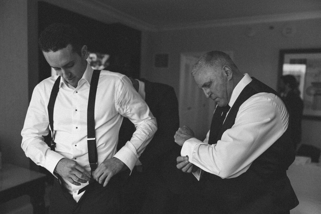 Candid moment of groom and groomsman adjusting formal attire indoors in black and white before wedding ceremony.