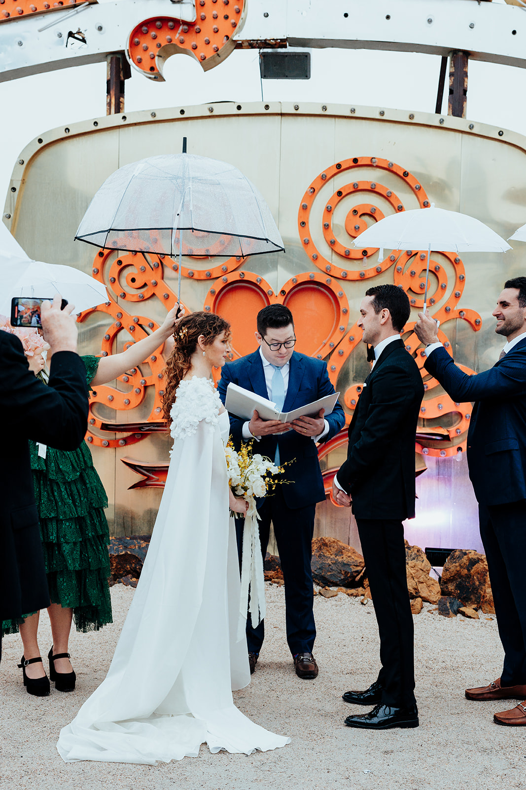 Bride and groom exchange vows during an outdoor wedding ceremony at the Neon Museum in Las Vegas, with vibrant vintage signage and umbrellas adding charm to the rainy day.