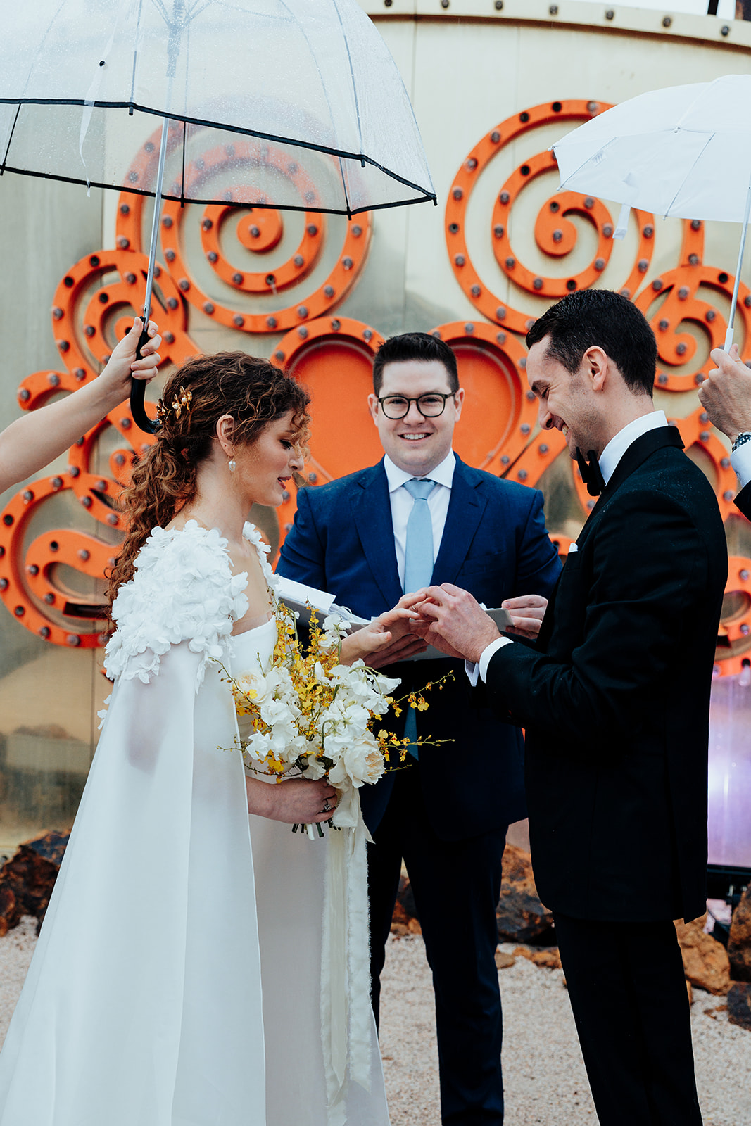 Bride and groom exchanging wedding rings during an outdoor ceremony at the Neon Museum in Las Vegas, surrounded by vintage signs and holding umbrellas in the rain.