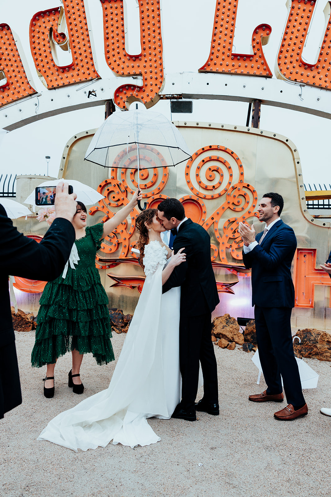 Bride and groom kissing during a vibrant wedding ceremony at the Neon Museum in Las Vegas, surrounded by neon signs and holding umbrellas on a rainy day.