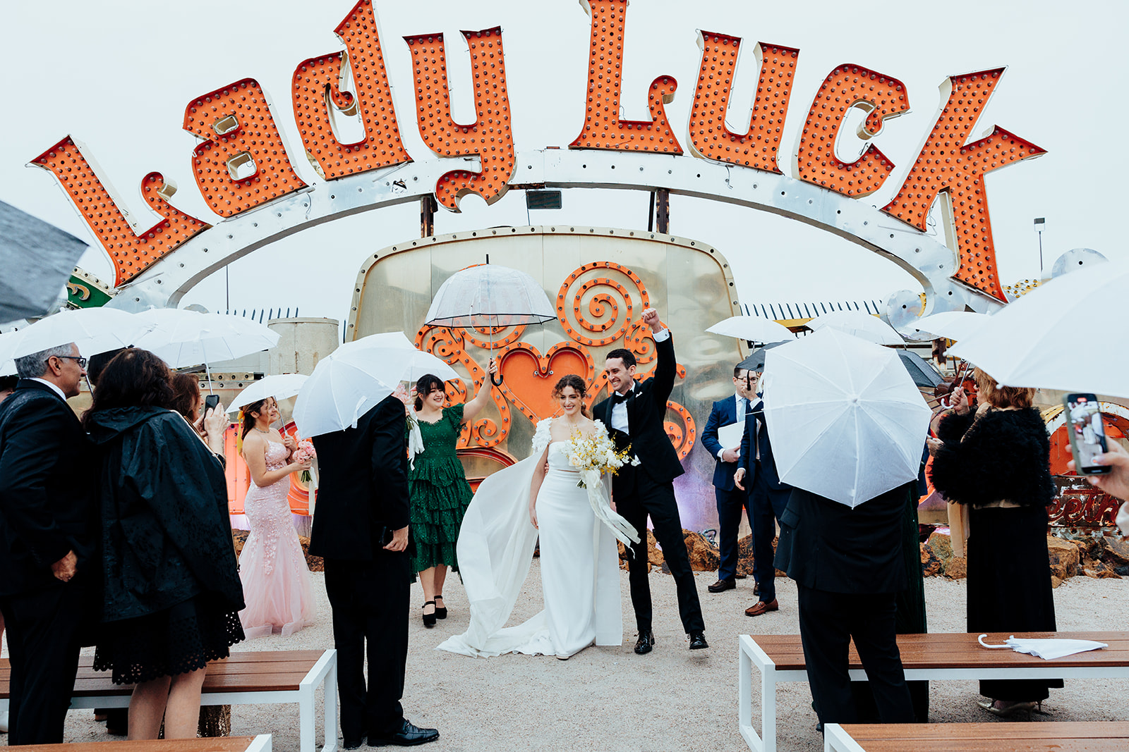 Bride and groom celebrating after their wedding ceremony at the Neon Museum in Las Vegas, surrounded by guests holding clear umbrellas under the "Lady Luck" neon sign.