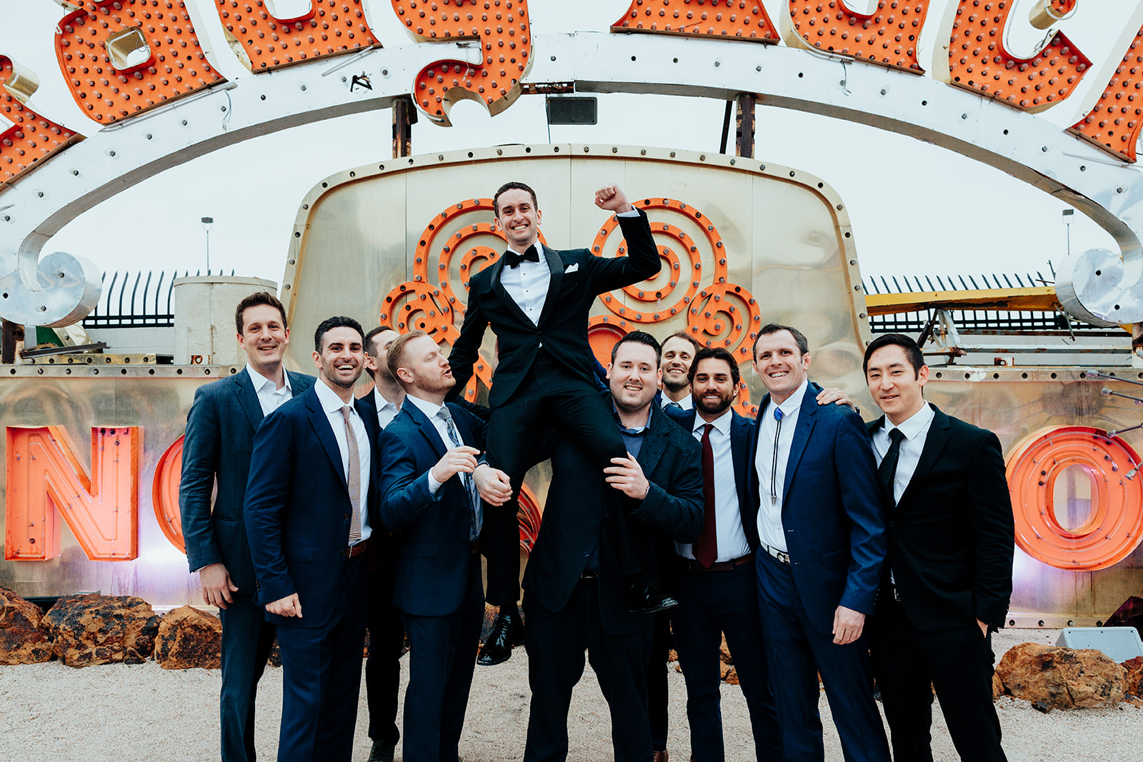 Groom in a tuxedo is lifted triumphantly by his groomsmen in front of the iconic "Lady Luck" neon sign at the Neon Museum in Las Vegas.