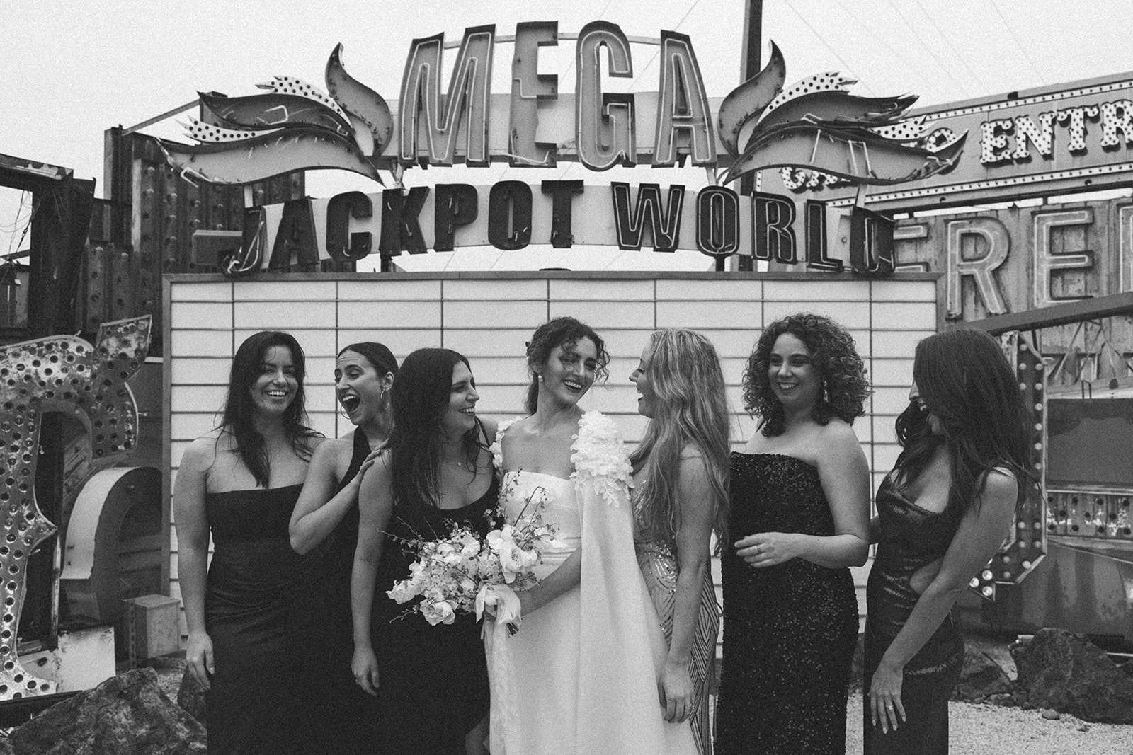 Black and white photo of a bride with her bridesmaids laughing together in front of the "Mega Jackpot World" sign at the Neon Museum in Las Vegas.