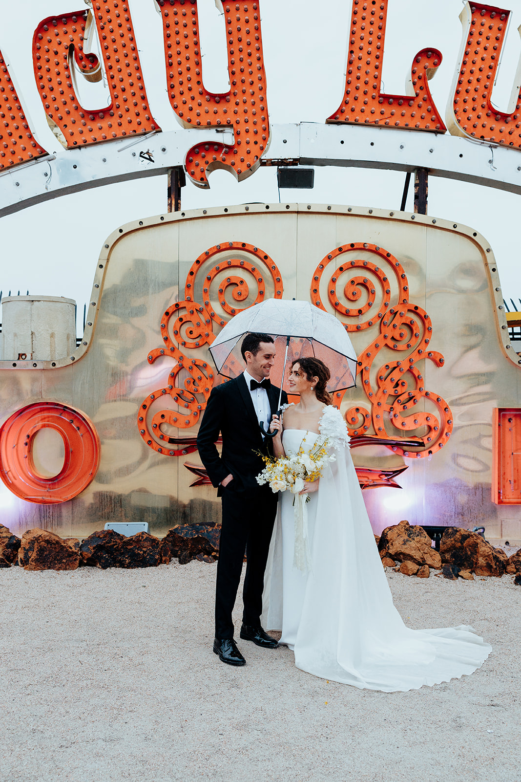 Newlywed couple standing under a transparent umbrella, with the bride holding a bouquet of white and yellow flowers, in front of a "Lady Luck" neon sign backdrop.