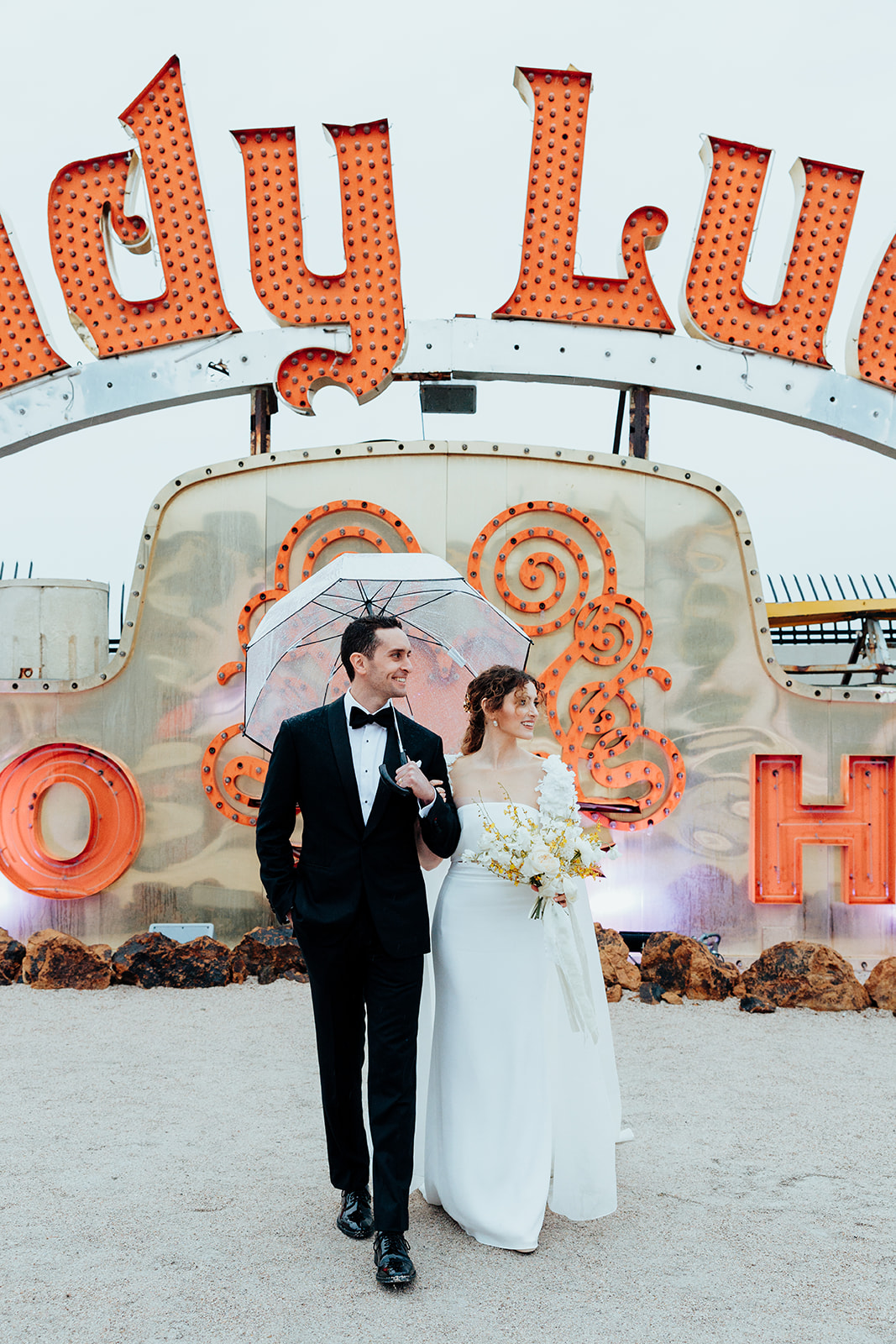 The bride and groom walking arm in arm under a clear umbrella, surrounded by vibrant neon signs, with the bride holding a bouquet of white and yellow flowers.