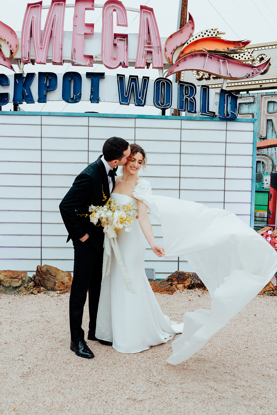 The groom kisses the bride on the cheek as her wedding cape flows in the breeze, standing before a vintage "Mega Jackpot World" neon sign. The bride holds a bouquet of white and yellow flowers.