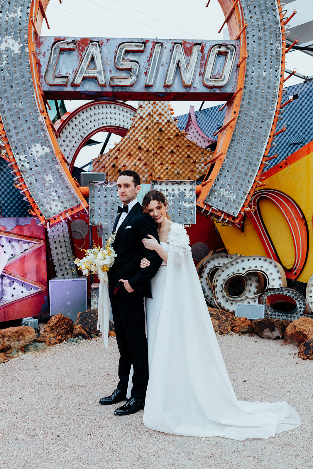 The bride leans on the groom with a warm smile as he stands confidently holding a bouquet of white and yellow flowers. They are framed by a vibrant retro "Casino" sign with colorful neon details in the background.