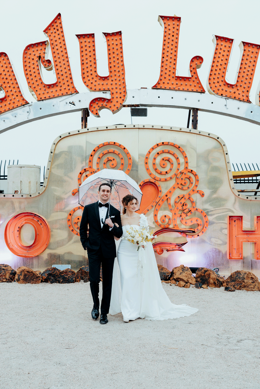 Newlywed couple standing under a transparent umbrella, with the bride holding a bouquet of white and yellow flowers, in front of a vibrant "Lady Luck" neon sign backdrop.