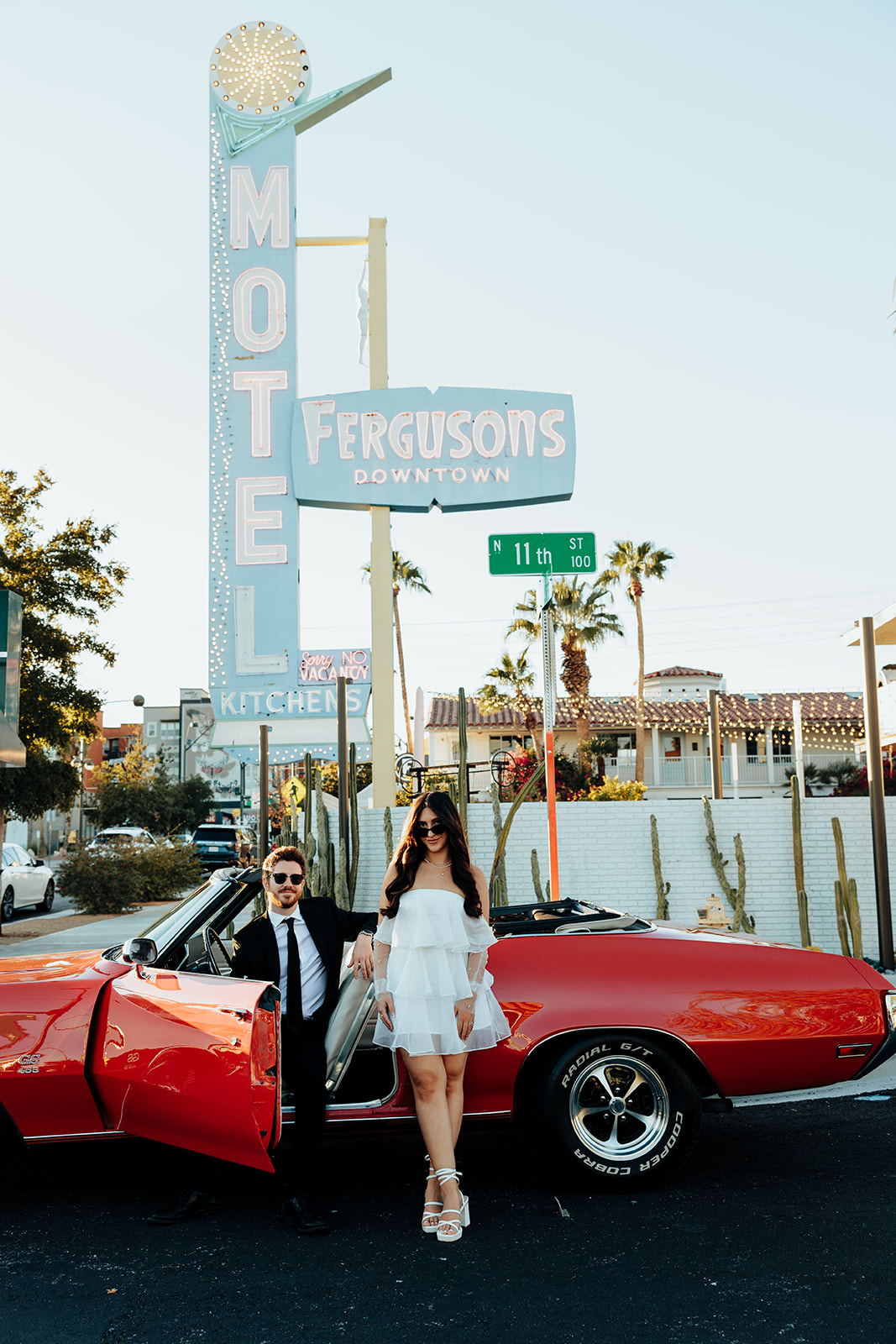 Engaged couple posing with a red vintage convertible under a retro motel sign, with the groom leaning on the car door and the bride standing beside him.