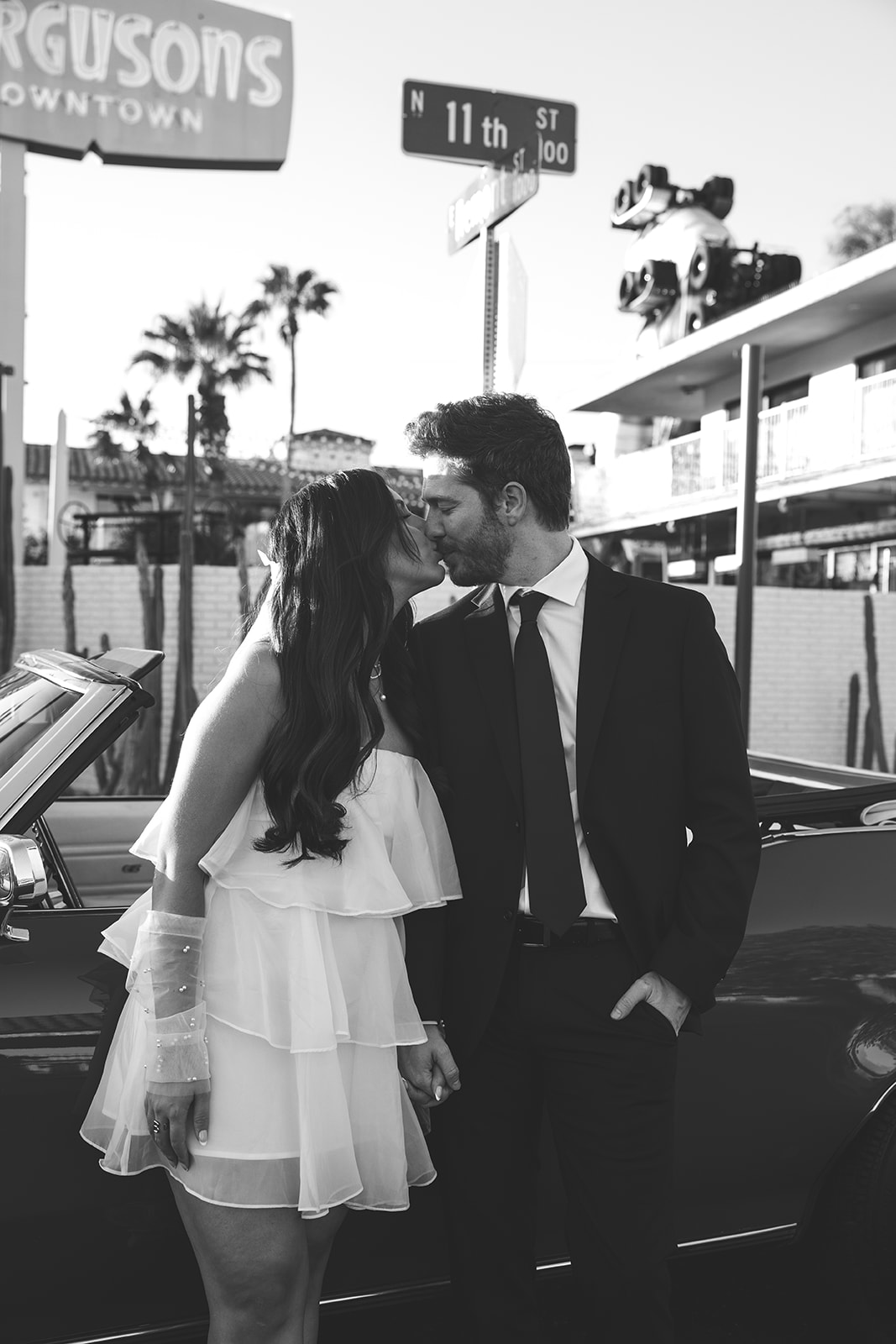 Black and white photo of an engaged couple sharing a romantic kiss in front of a vintage motel sign and red convertible.