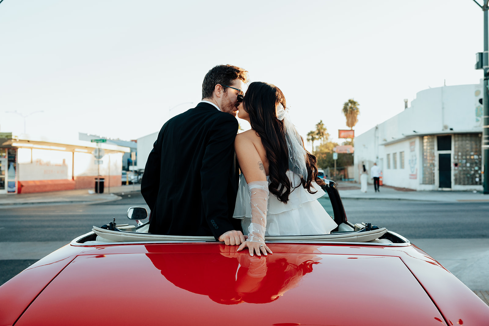 Engaged couple sitting on the back of a red vintage convertible, sharing a kiss as the sun sets.