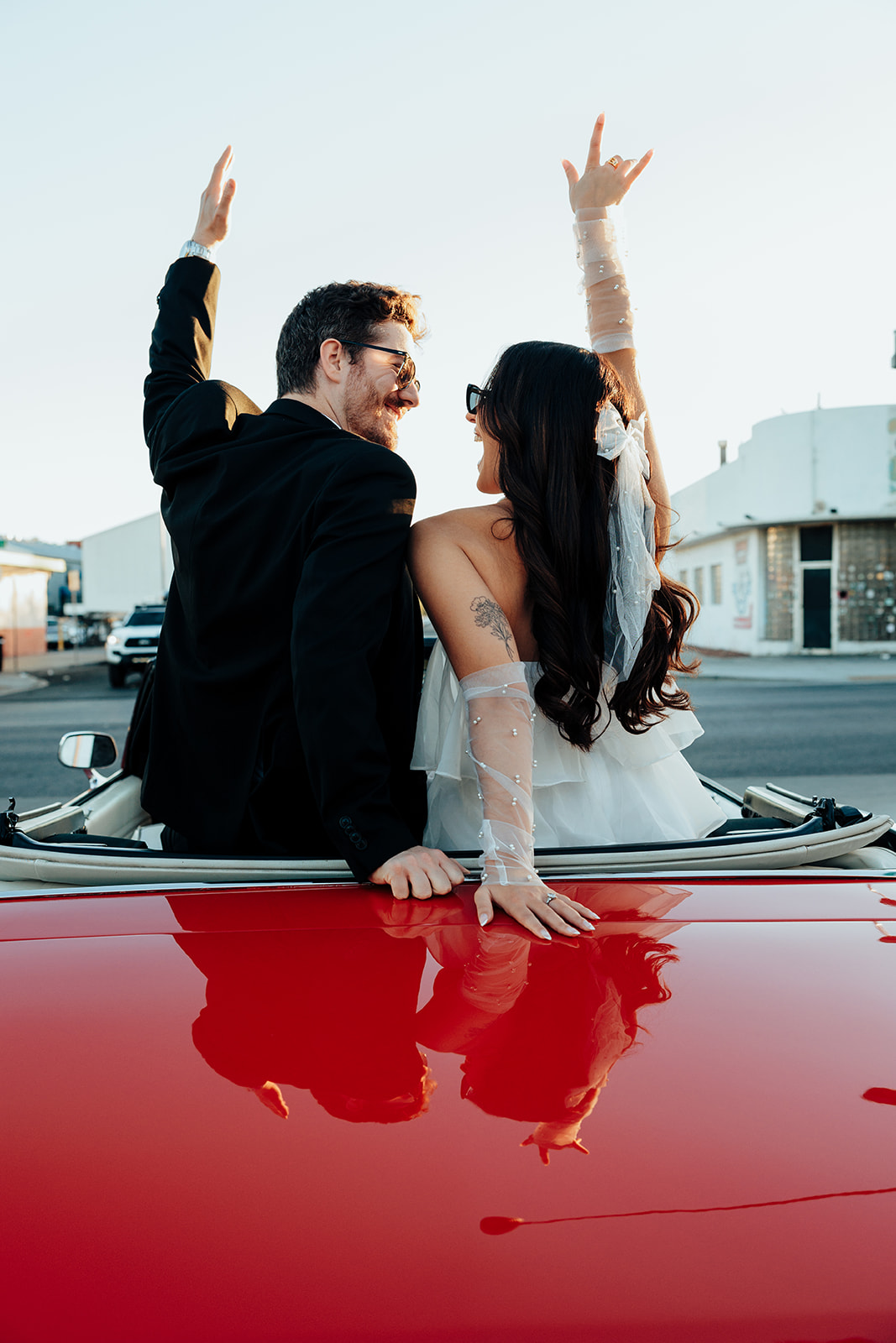Engaged couple sitting on the back of a red vintage convertible, laughing and raising their hands in excitement at sunset.