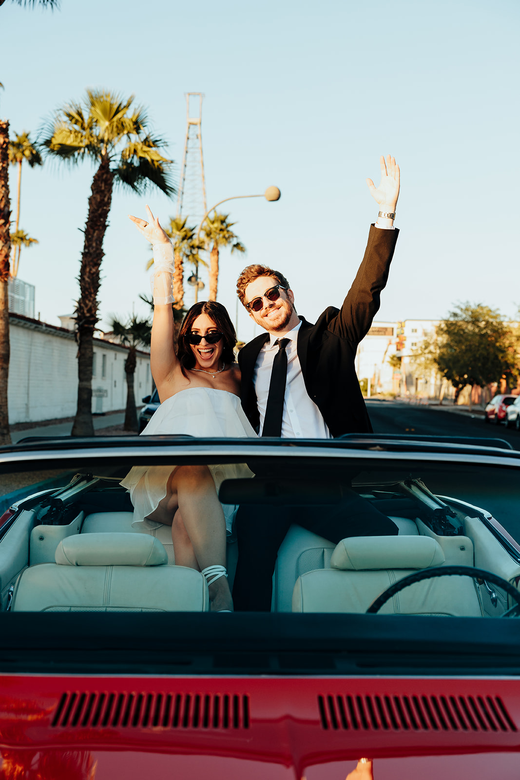 Engaged couple standing in a red convertible, smiling and cheering with their hands raised under the palm trees at sunset.