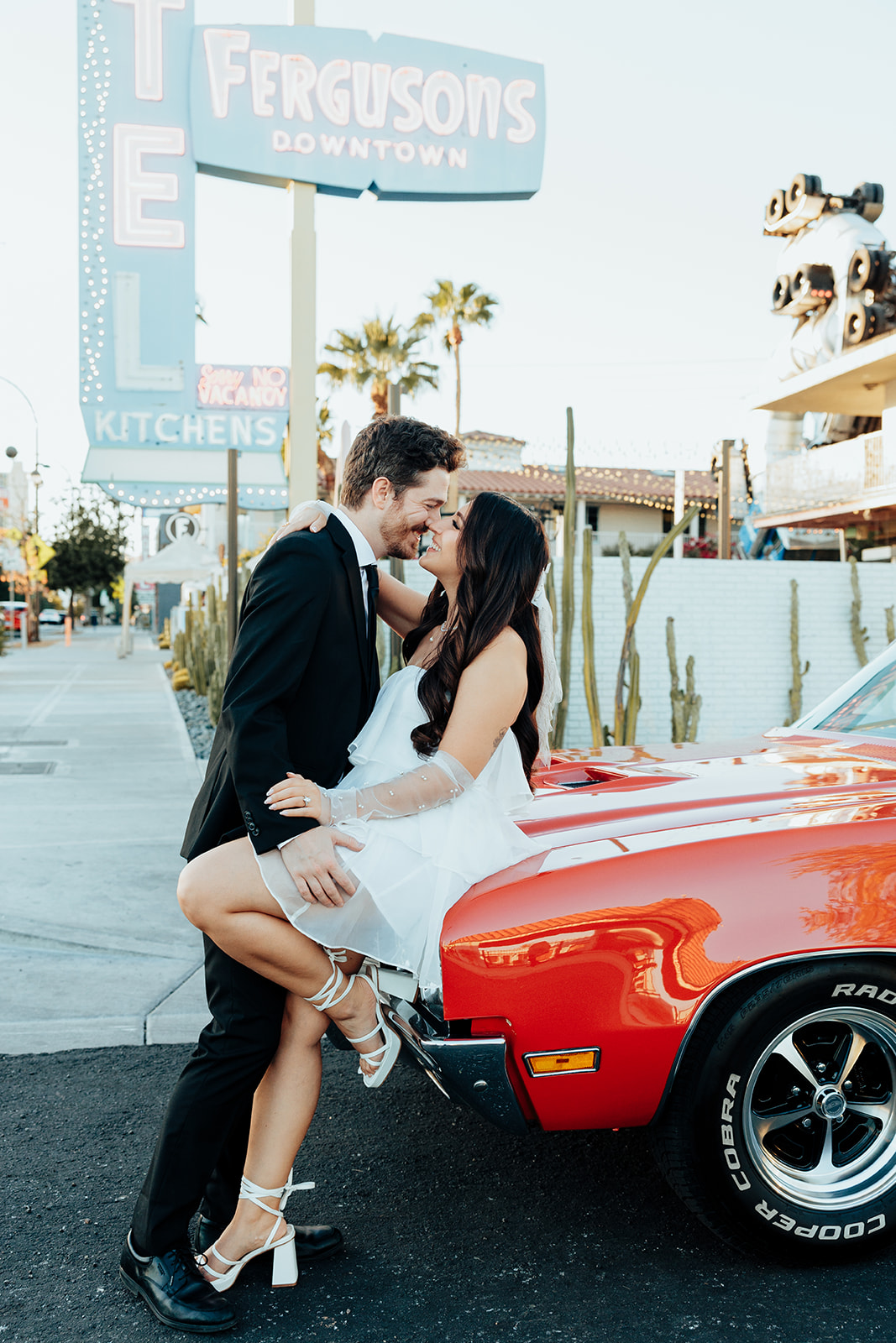Engaged couple sharing a romantic moment while leaning against a red vintage convertible under a retro motel sign.
