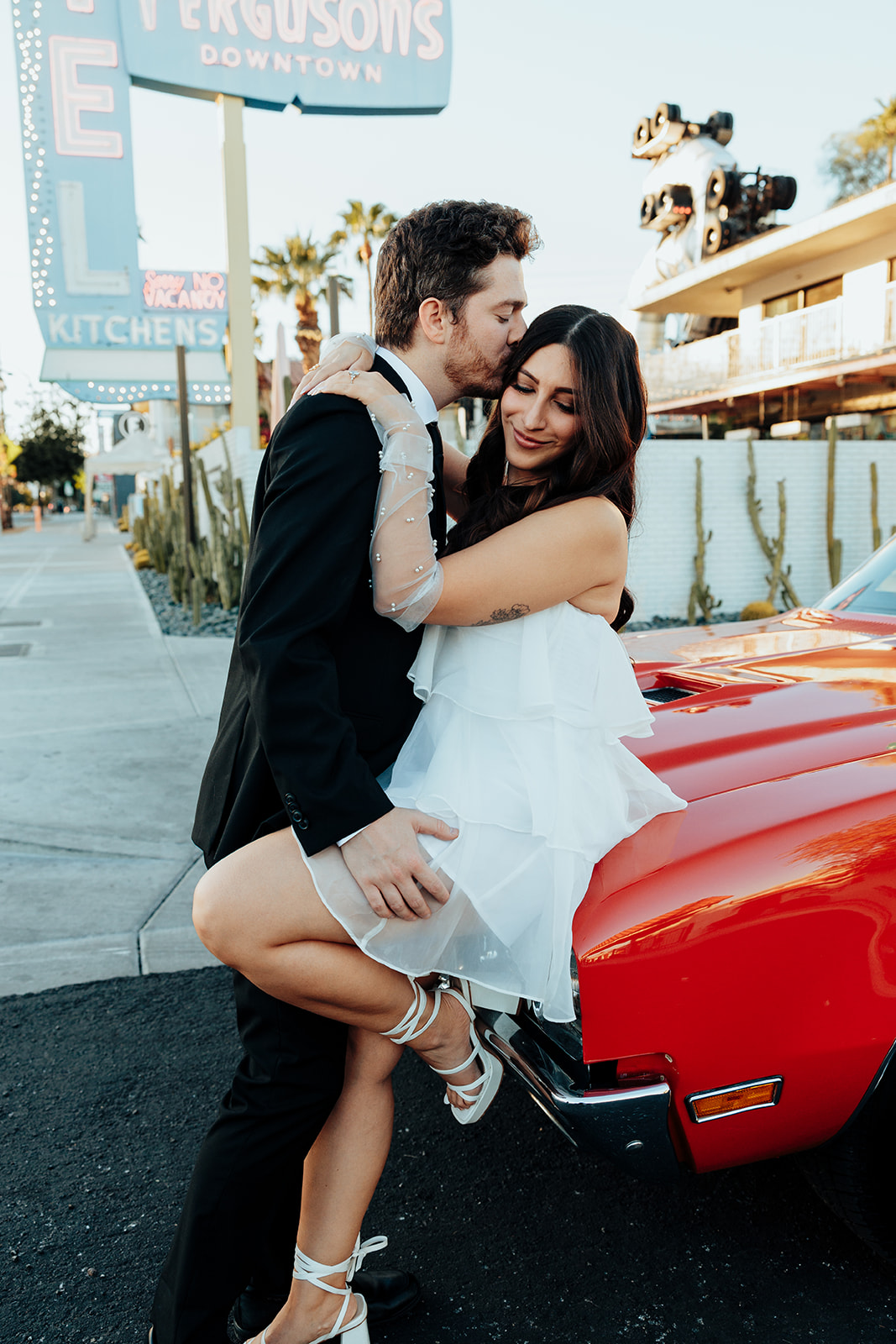 Engaged couple sharing a romantic embrace against a red vintage convertible, with the groom kissing the bride’s forehead.
