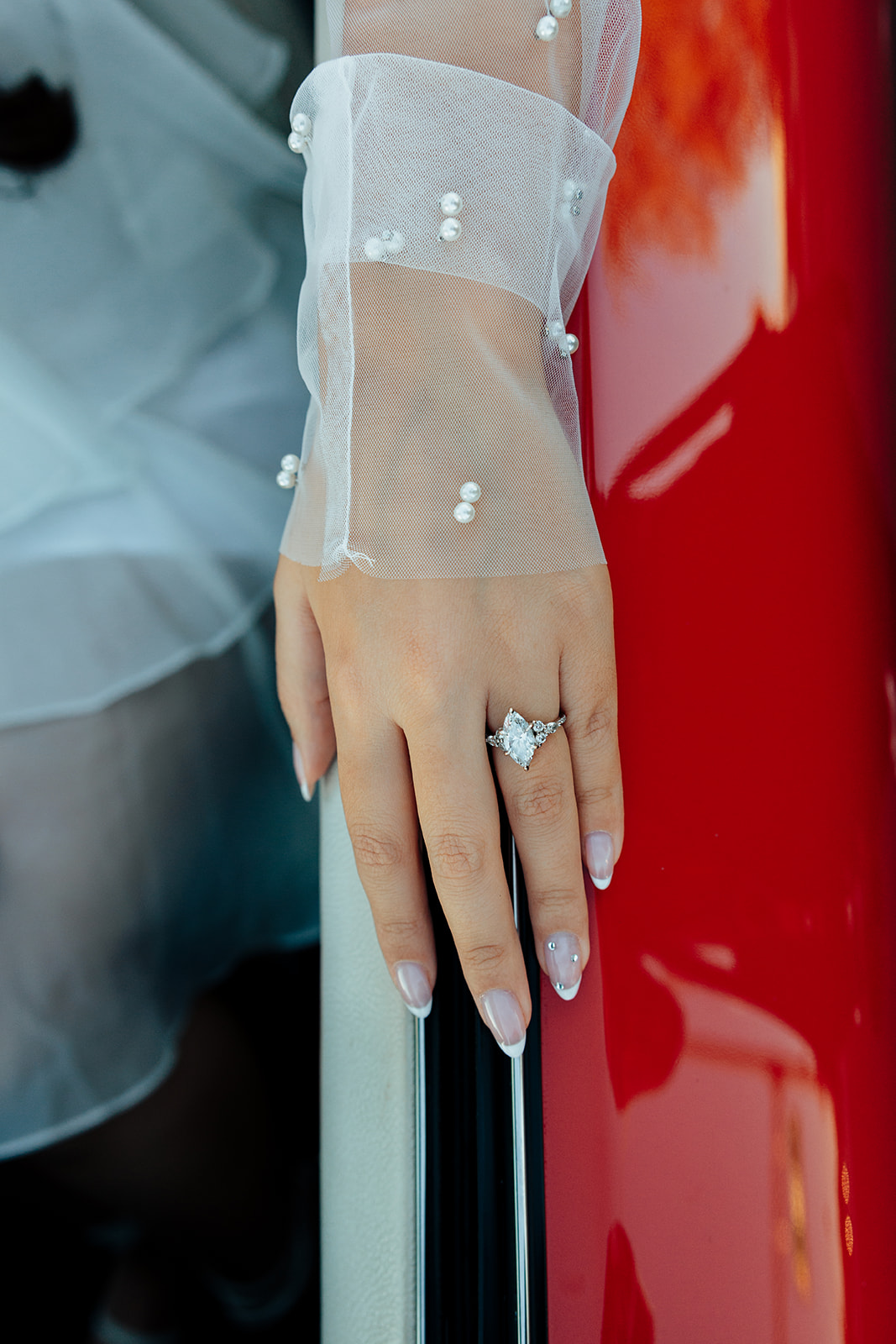 Close-up of a bride’s hand resting on a red convertible, showcasing her elegant engagement ring with a pear-cut diamond.