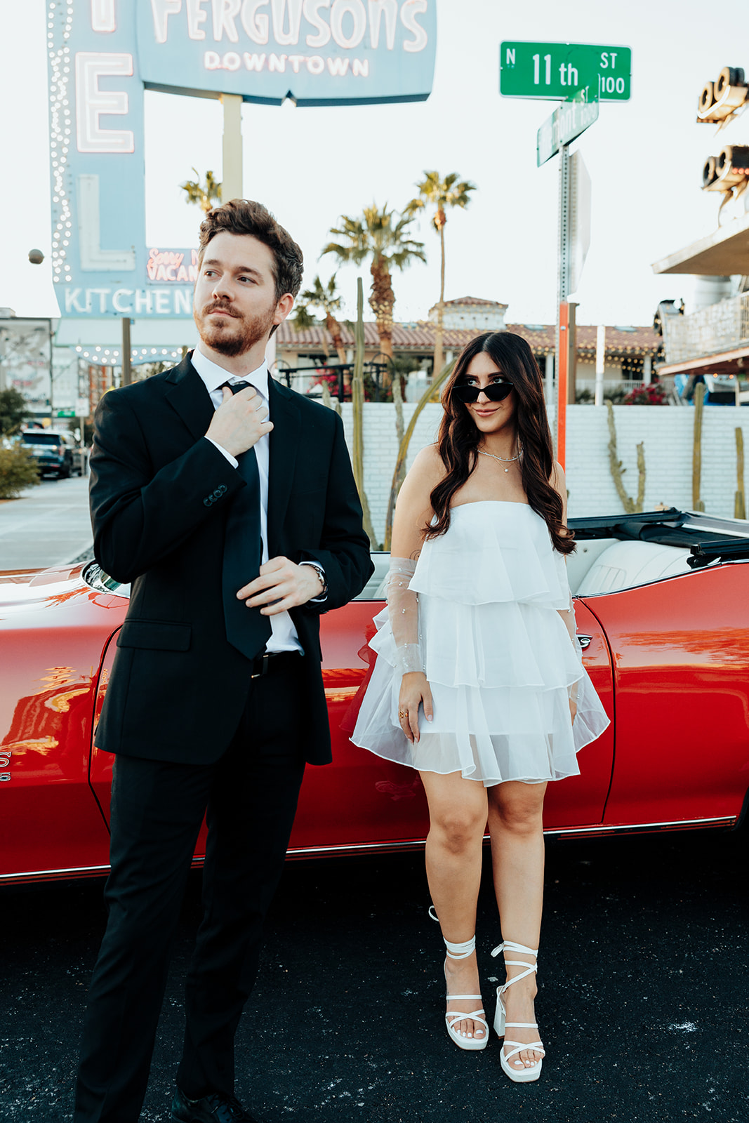 Engaged couple posing confidently in front of a red vintage convertible, with the groom adjusting his tie and the bride smiling in sunglasses.