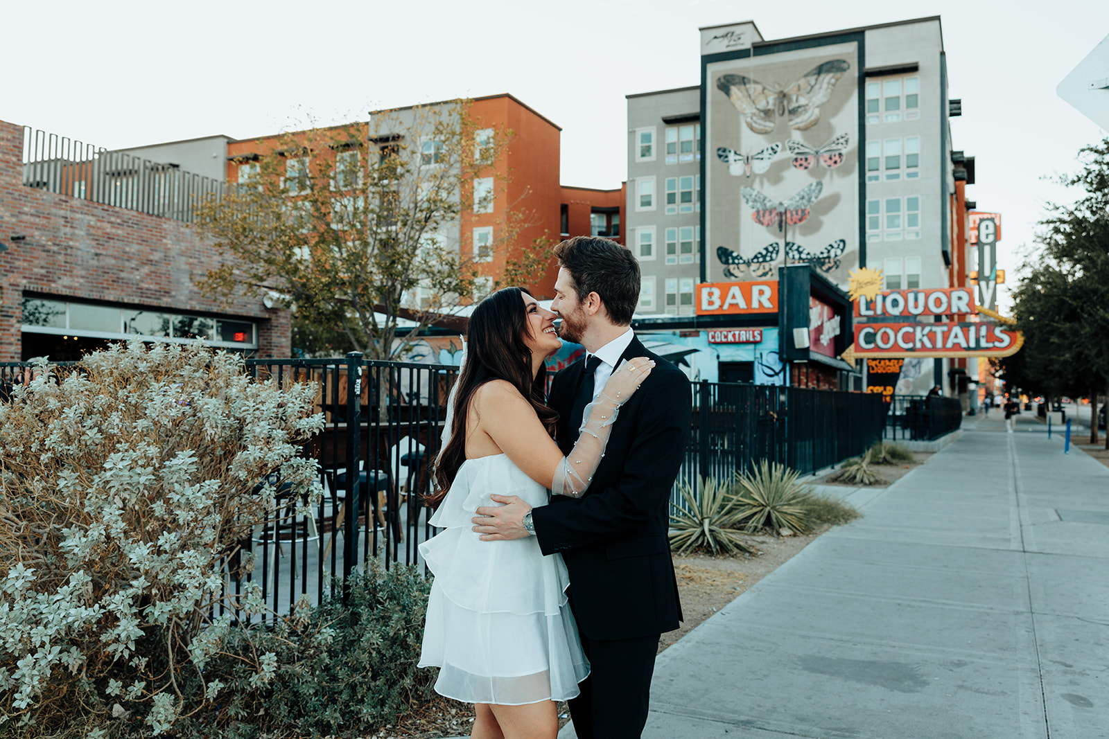 Engaged couple sharing a joyful embrace on a city sidewalk, with colorful buildings and neon bar signs in the background.