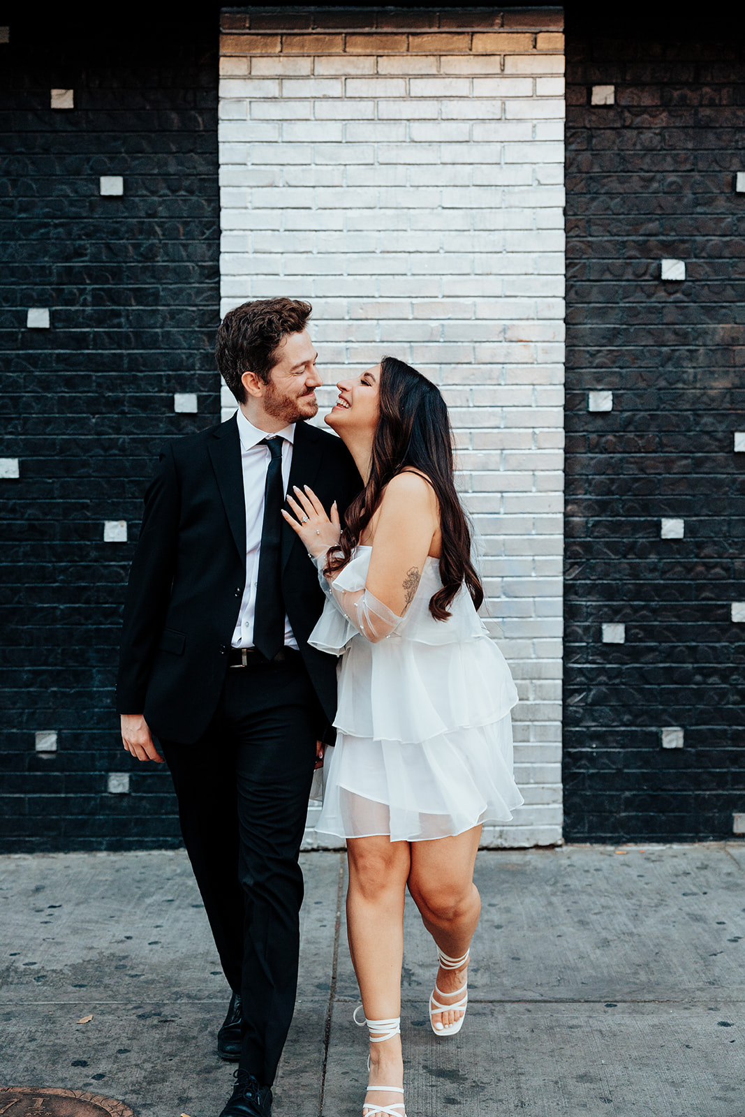 Engaged couple laughing and walking hand in hand against a modern black-and-white brick wall.