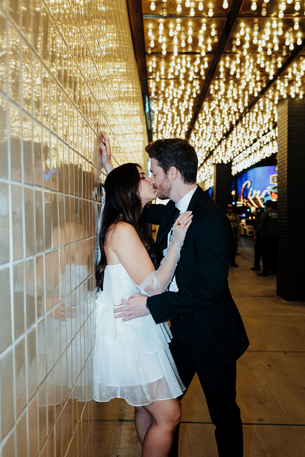 Engaged couple sharing a romantic kiss under glowing marquee lights, with the bride leaning against a tiled wall.