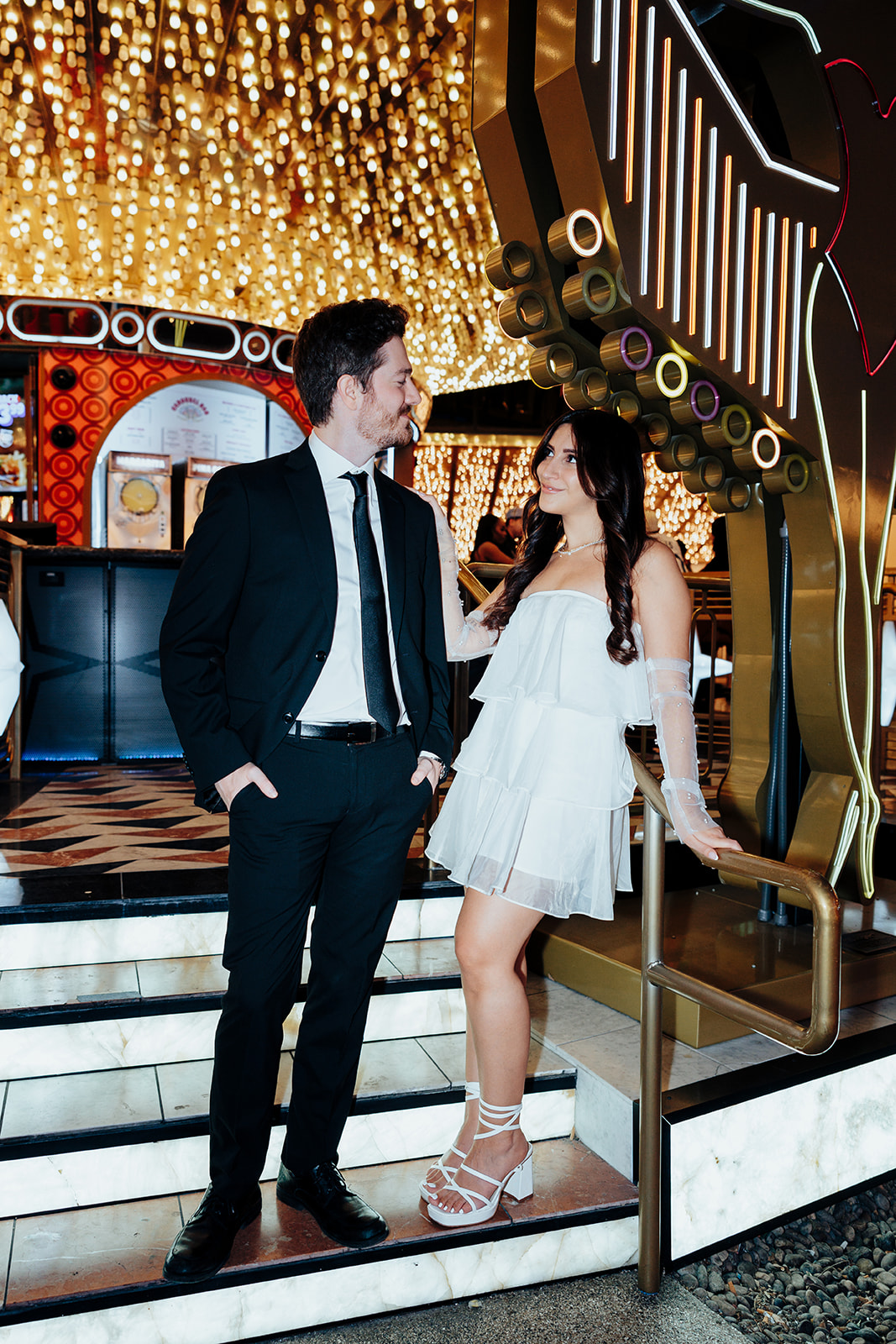 Couple standing on elegant staircase under bright marquee lights, dressed in stylish engagement attire.