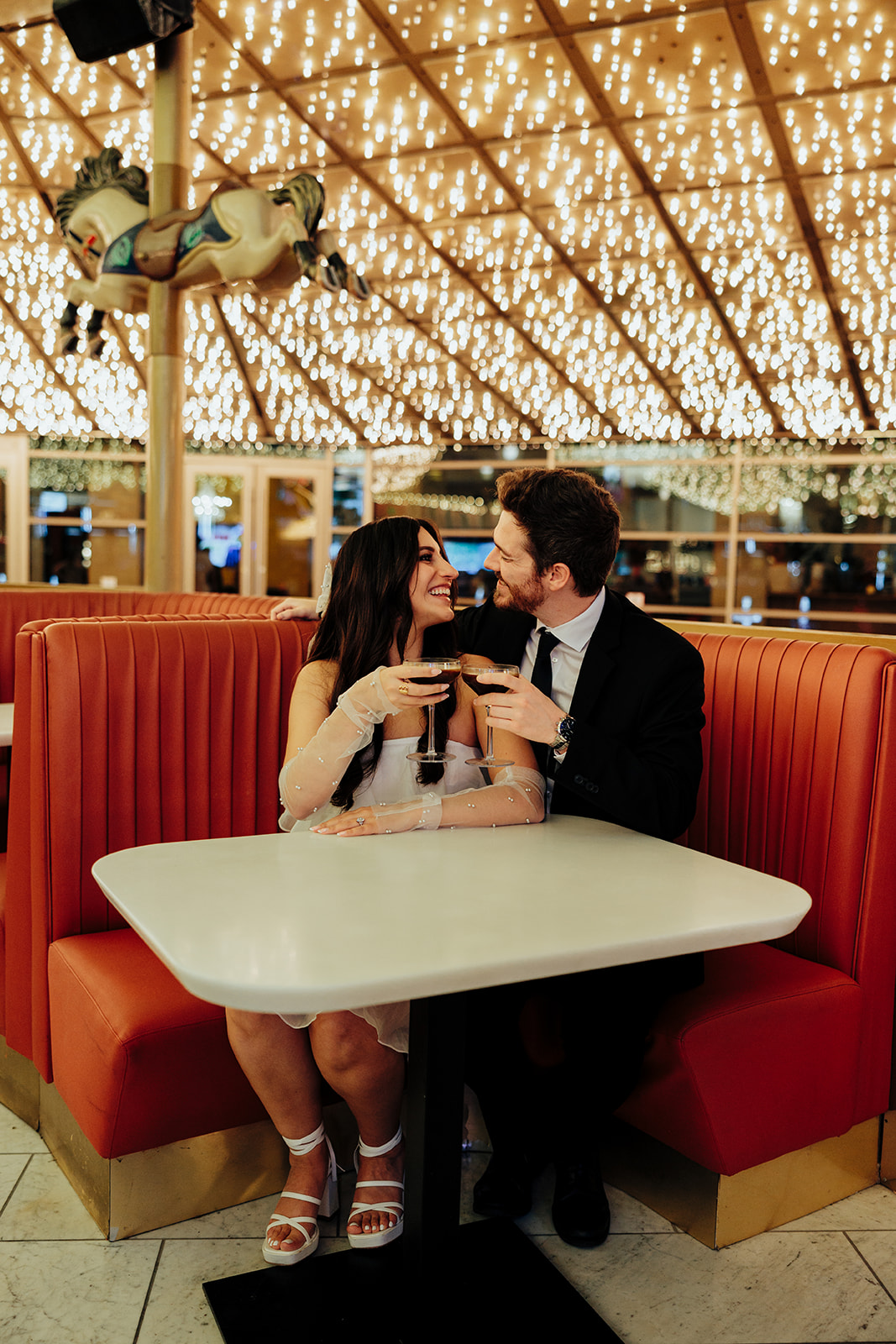 Engaged couple toasting with espresso martinis in a retro diner booth, smiling at each other under twinkling lights.