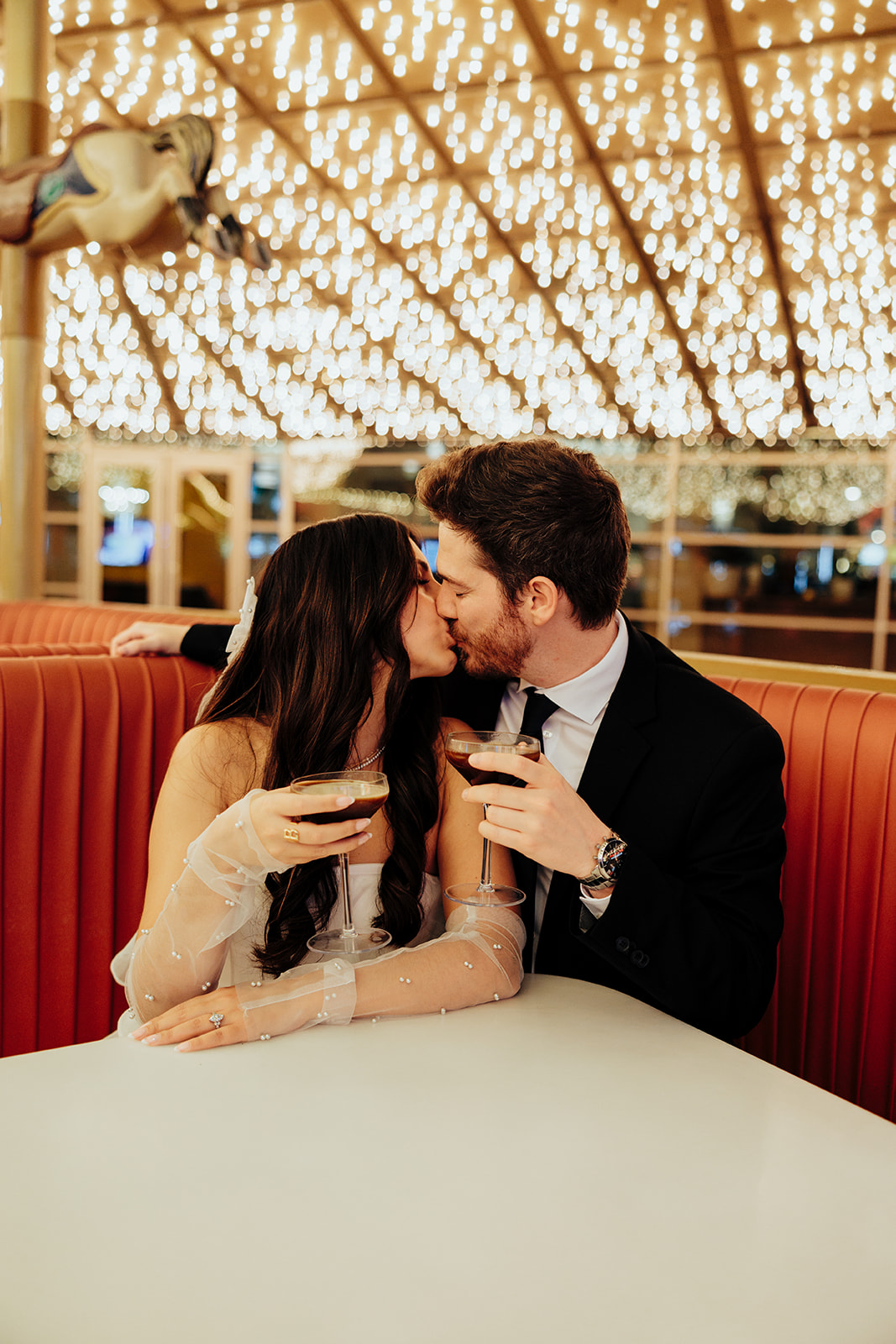 Couple sharing a kiss while holding espresso martinis, with a glowing ceiling of string lights in the background.
