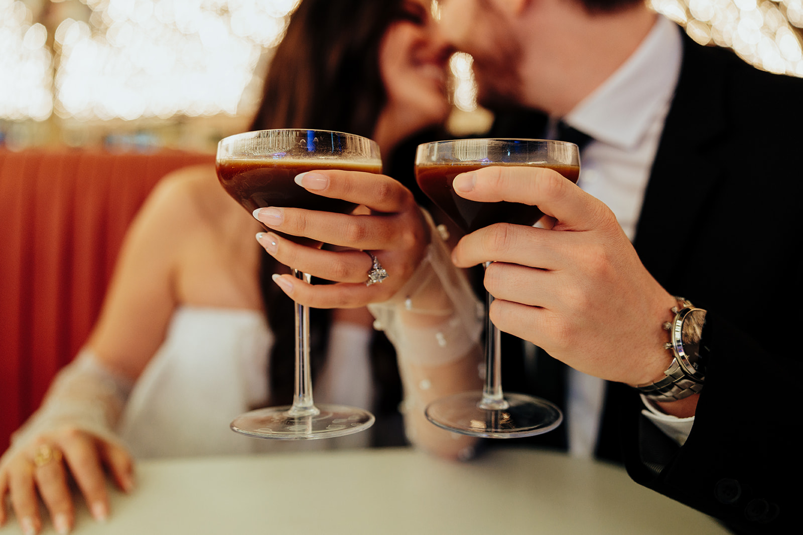 Close-up of a couple's hands holding espresso martini glasses, with the engagement ring in focus.