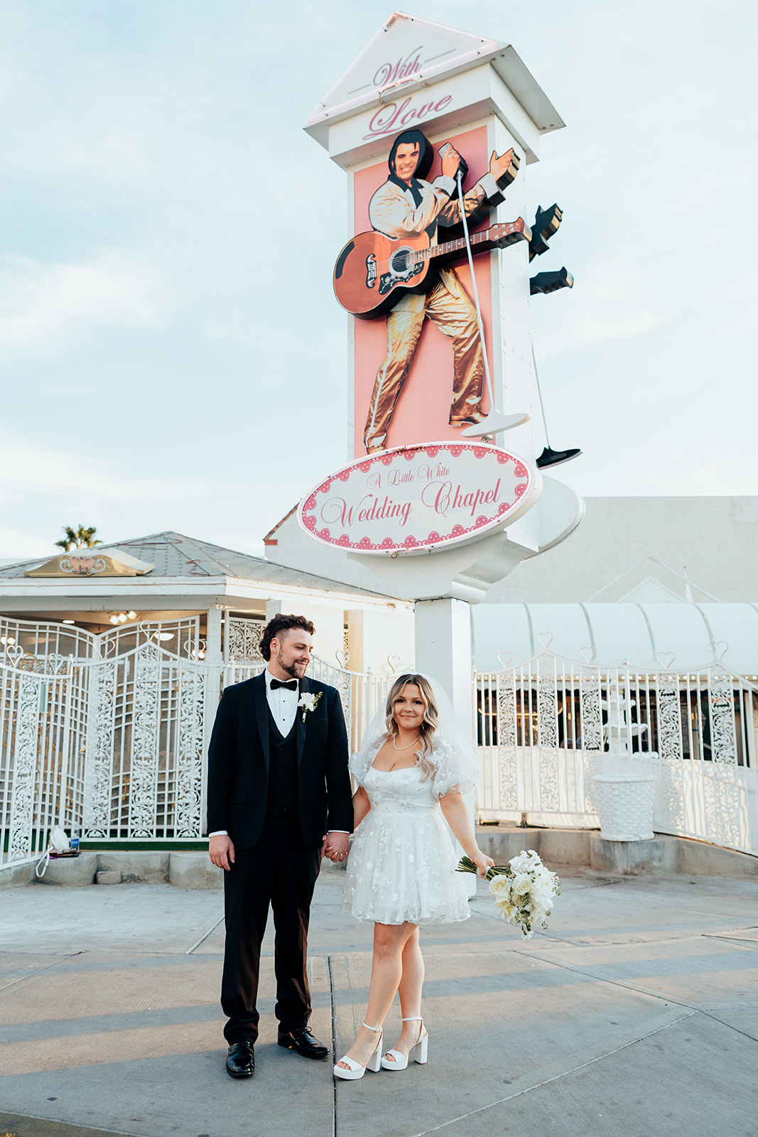 Bride and groom posing for an elopement photo at Little White Wedding Chapel, with the Elvis Presley sign and floral decor in the background.