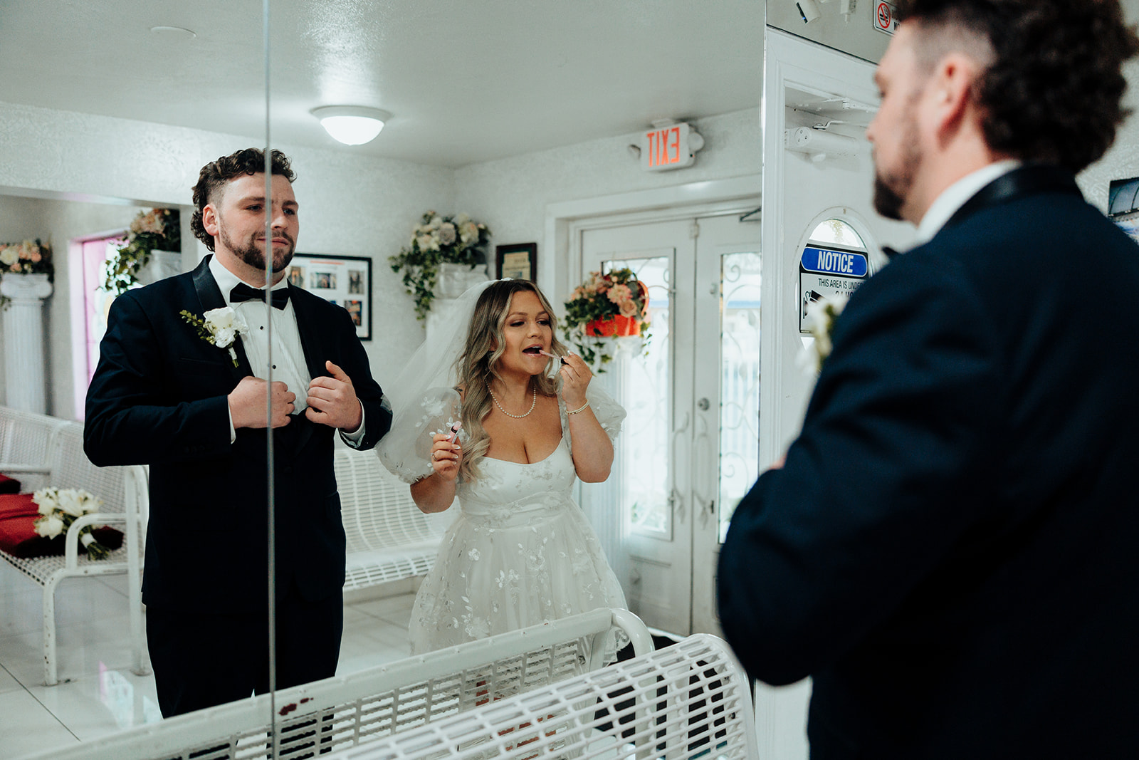 Bride applying makeup and groom adjusting tie in a mirror at Little White Wedding Chapel elopement, with white benches and floral details.