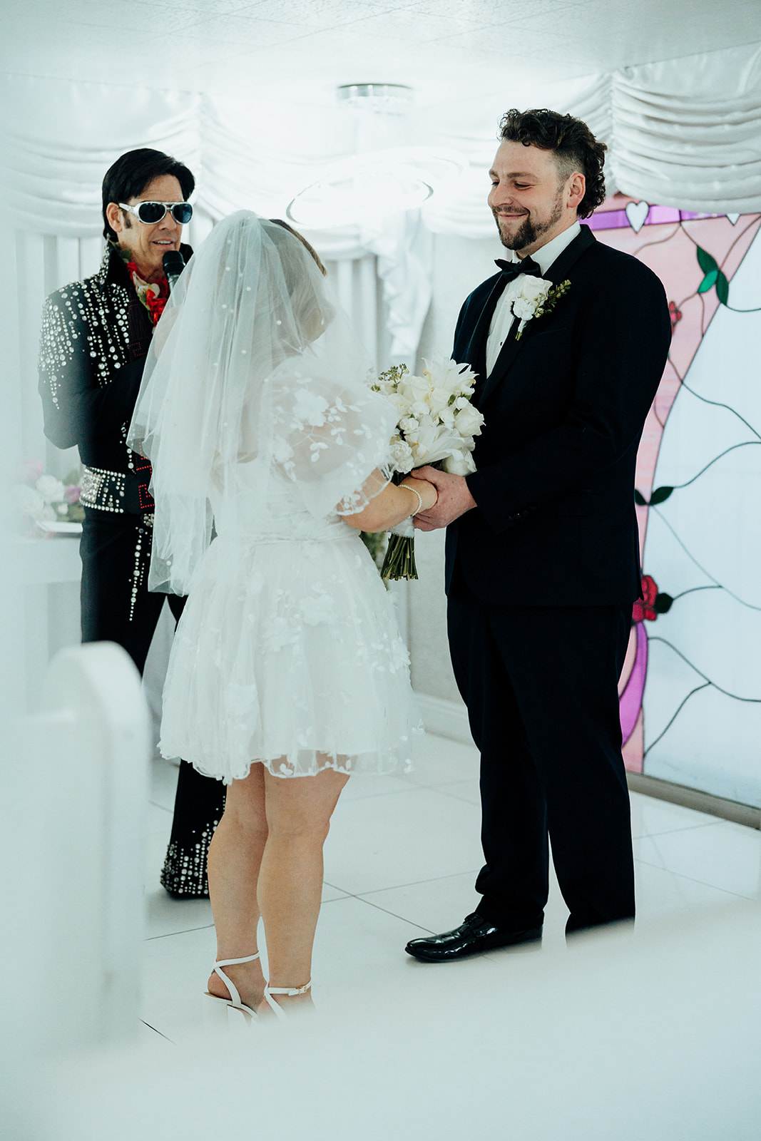 Bride and groom exchanging vows at Little White Wedding Chapel elopement, officiated by Elvis impersonator, with white drapes and stained-glass window in the background.