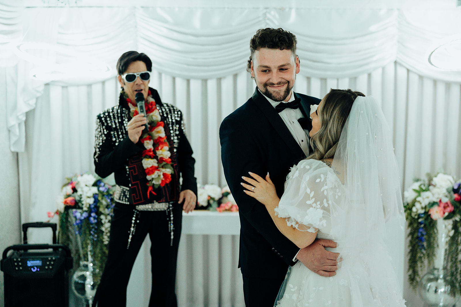 Bride and groom embracing during elopement at Little White Wedding Chapel, with Elvis impersonator officiating and floral decor in the background.