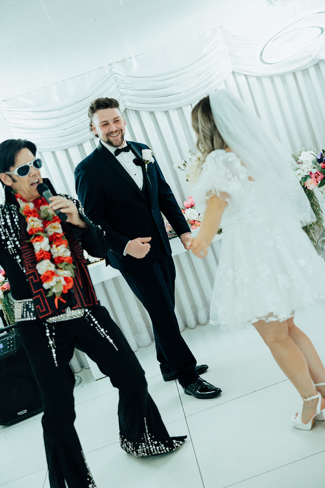 Elvis impersonator officiating an elopement at Little White Wedding Chapel with bride in white gown and groom in tuxedo, holding hands with floral decor in the background.